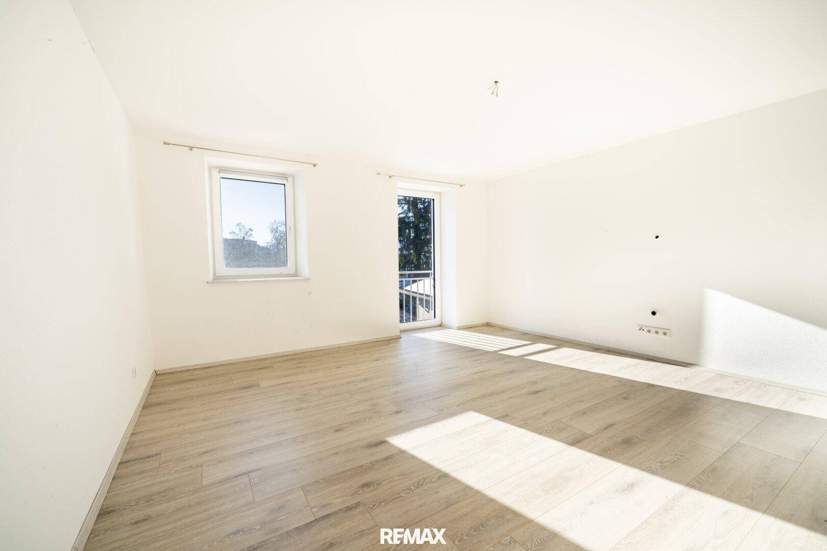 Lichtdurchfluteter Raum mit hellem Laminatboden, Fenster und Zugang zu einem französischen Balkon.