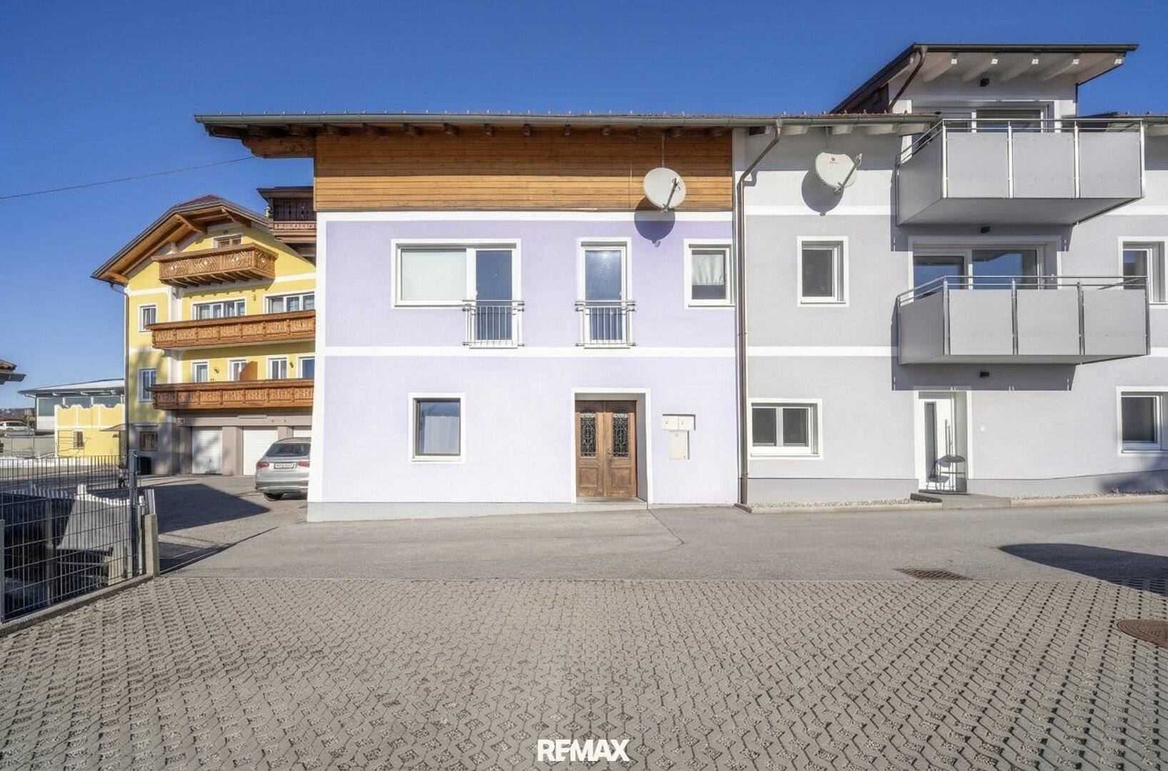 Außenansicht eines Gebäudes mit lila und grauer Fassade sowie gepflastertem Vorplatz.