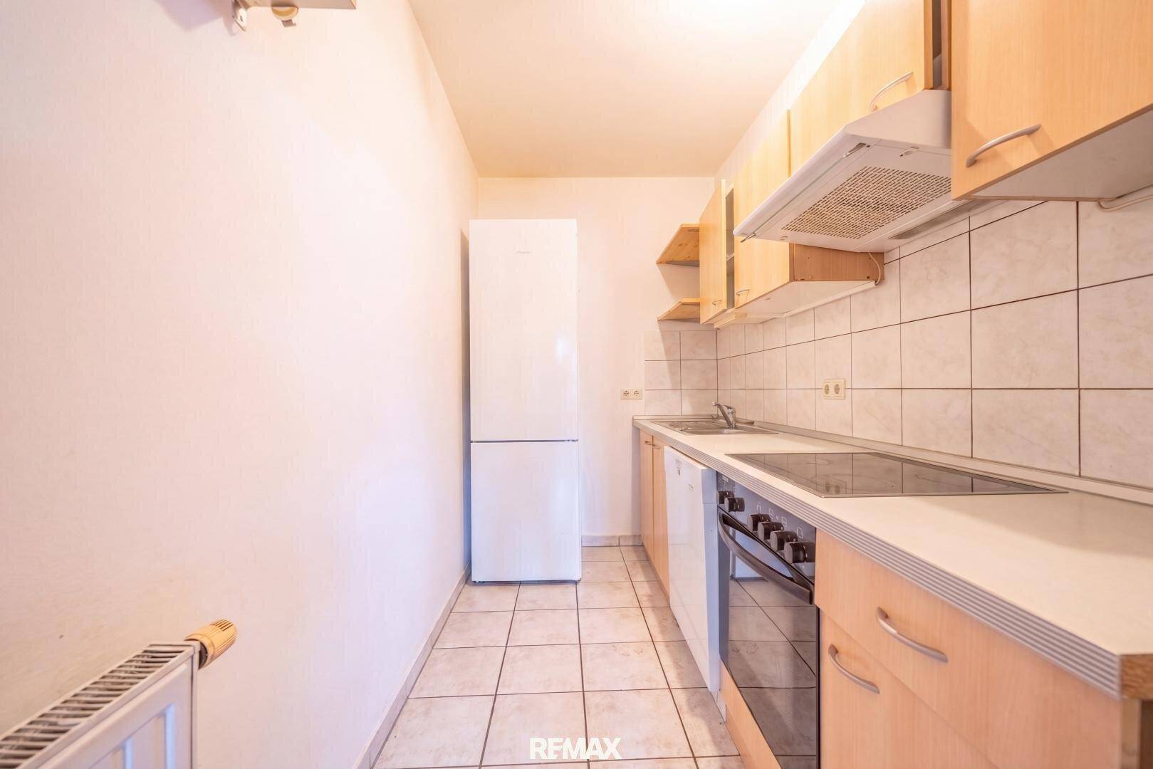 Kleine Küche mit hellen Holzschränken, Fliesenboden und weißem Kühlschrank.