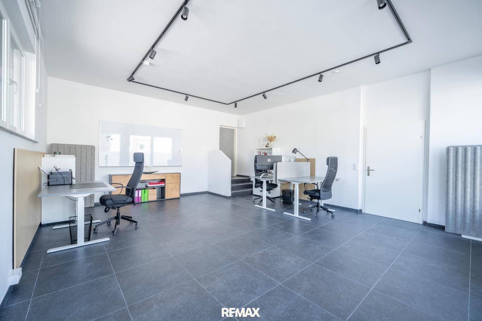 Geräumiges Büro mit mehreren Arbeitsplätzen, dunklen Fliesen und einem Treppenaufgang im Hintergrund.