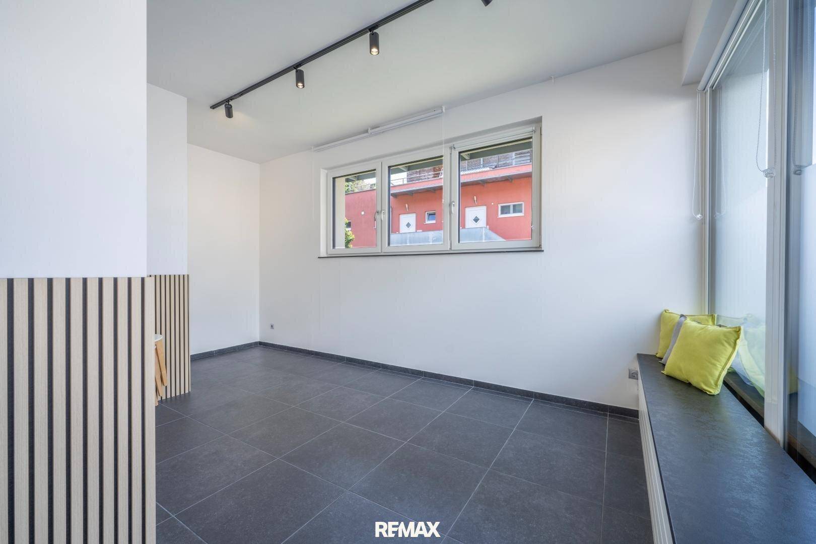 Geräumiger Büroraum mit dunklen Fliesen, großen Fenstern und einer Sitzbank mit Kissen.
