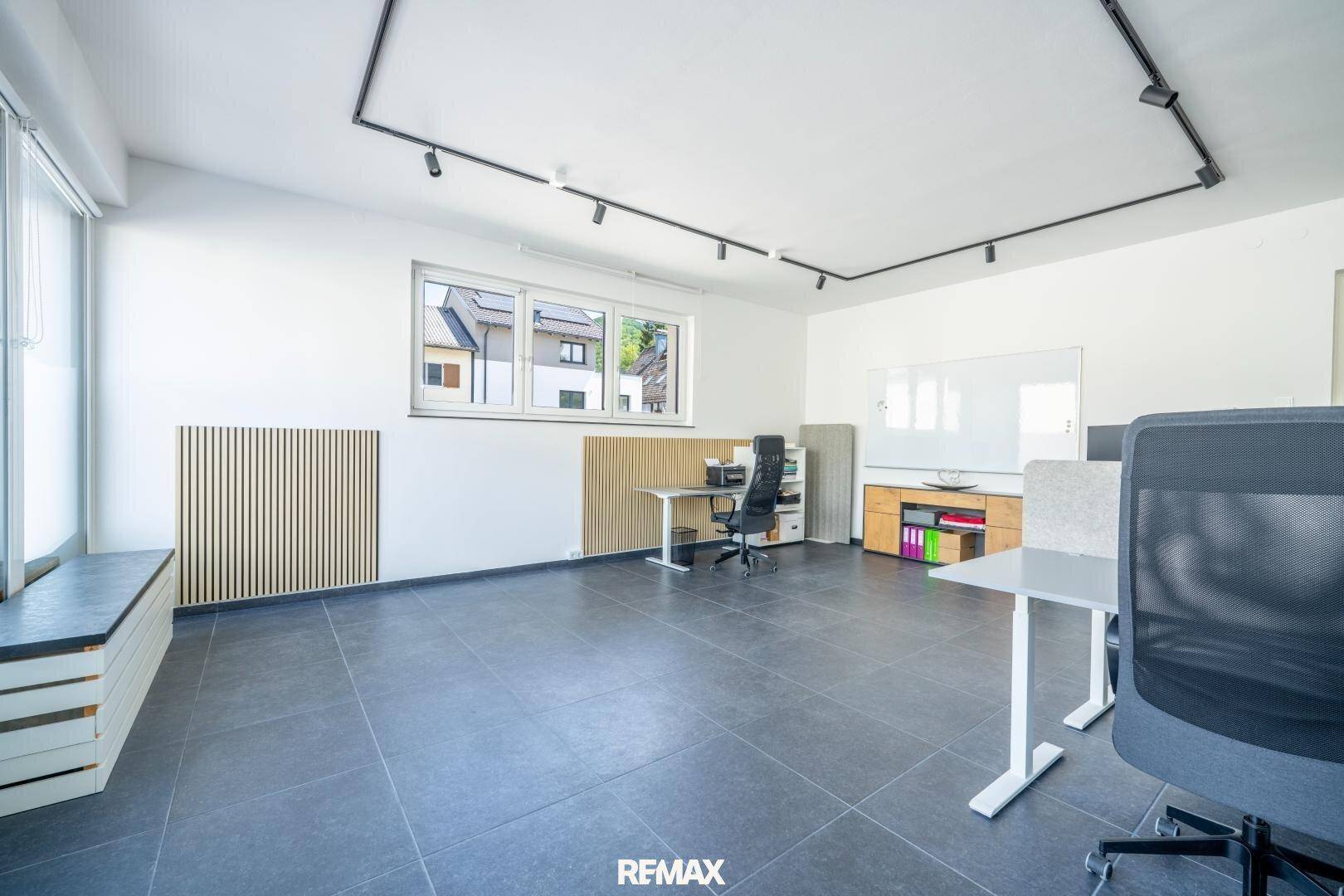Modern eingerichtetes Büro mit mehreren Arbeitsplätzen, dunklen Fliesen und großen Fenstern.