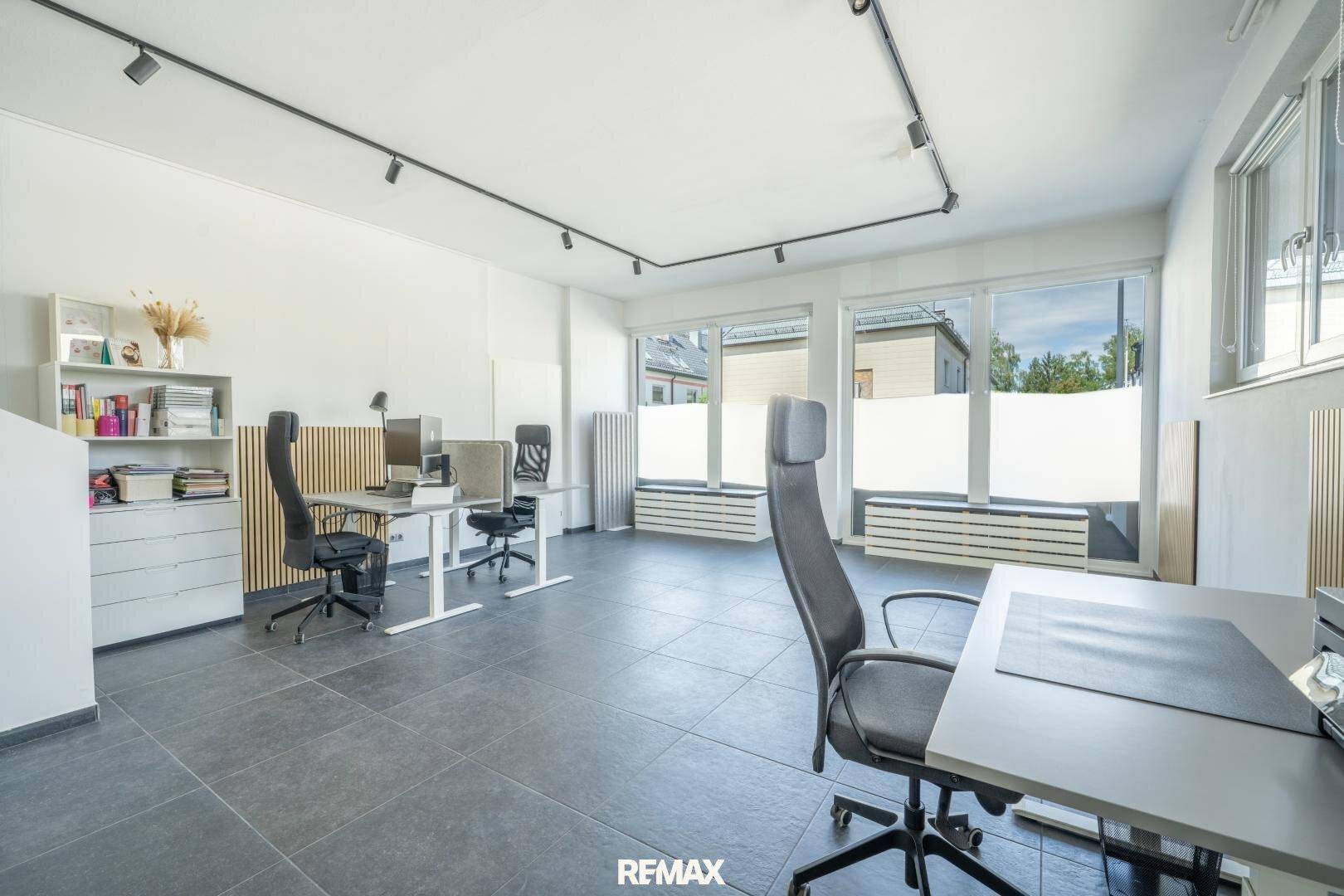 Modernes Büro mit mehreren Arbeitsplätzen, großen Fenstern und dunklen Fliesenboden.