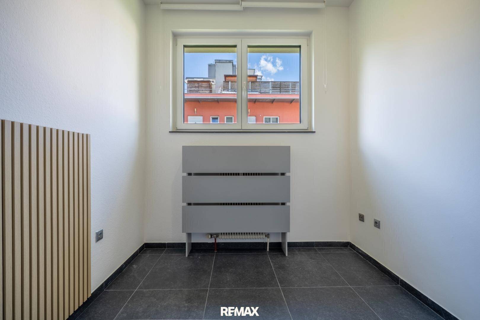 Kleiner Büroraum mit Fenster, dunklen Fliesen und einem modernen Heizkörper unter dem Fenster.