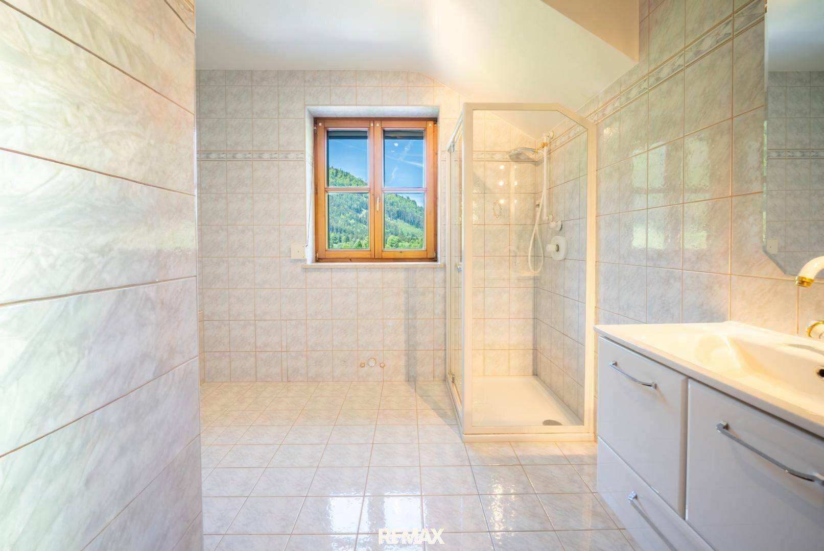 Gefliestes Badezimmer mit Duschkabine, Einzelwaschbecken und Fenster mit Blick ins Grüne.