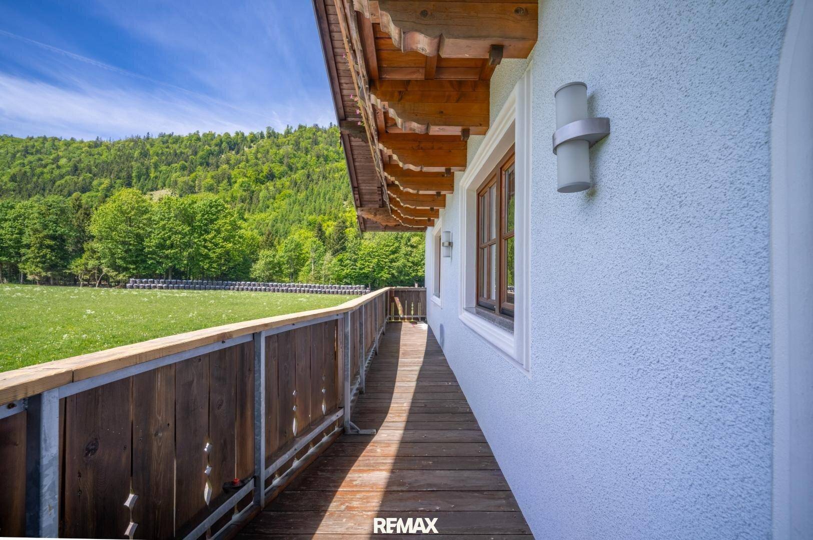 Langer Holzbalkon entlang des Hauses mit weitem Blick auf grüne Wiesen und bewaldete Hügel.