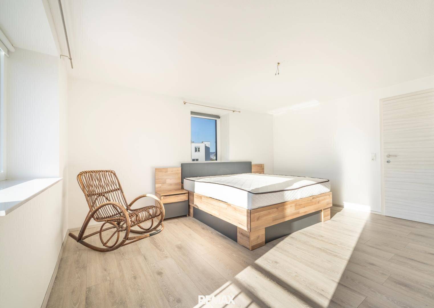 Helles Schlafzimmer mit Holzbett, Schaukelstuhl und viel Tageslicht durch das Fenster.