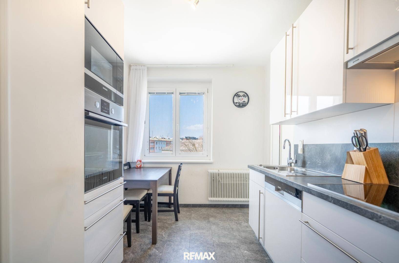 Moderne Küche mit weißen Schränken, integrierten Geräten und kleinem Essbereich am Fenster.