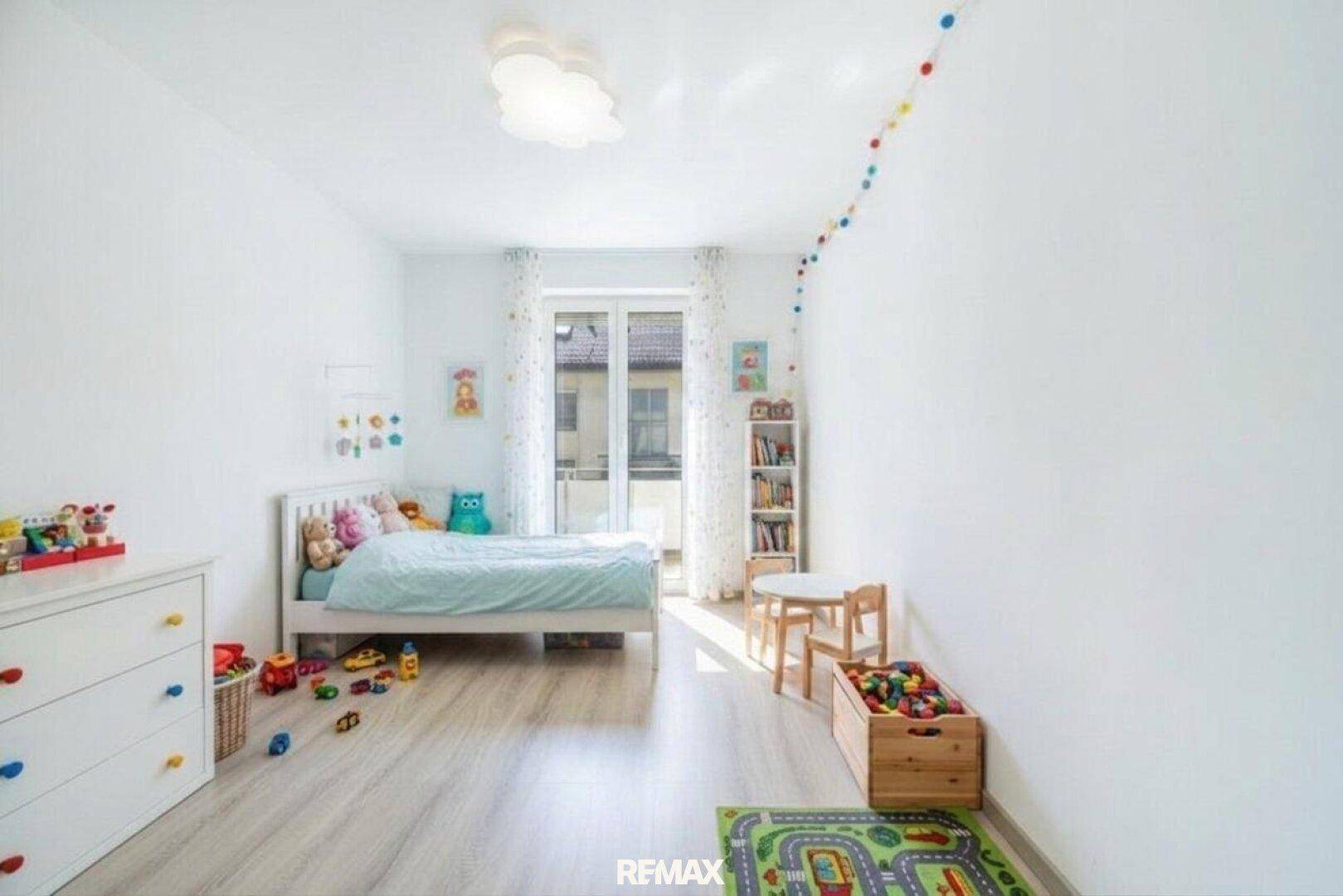 Helles Kinderzimmer mit Laminatboden, Spielzeug und Zugang zu einem kleinen Balkon.