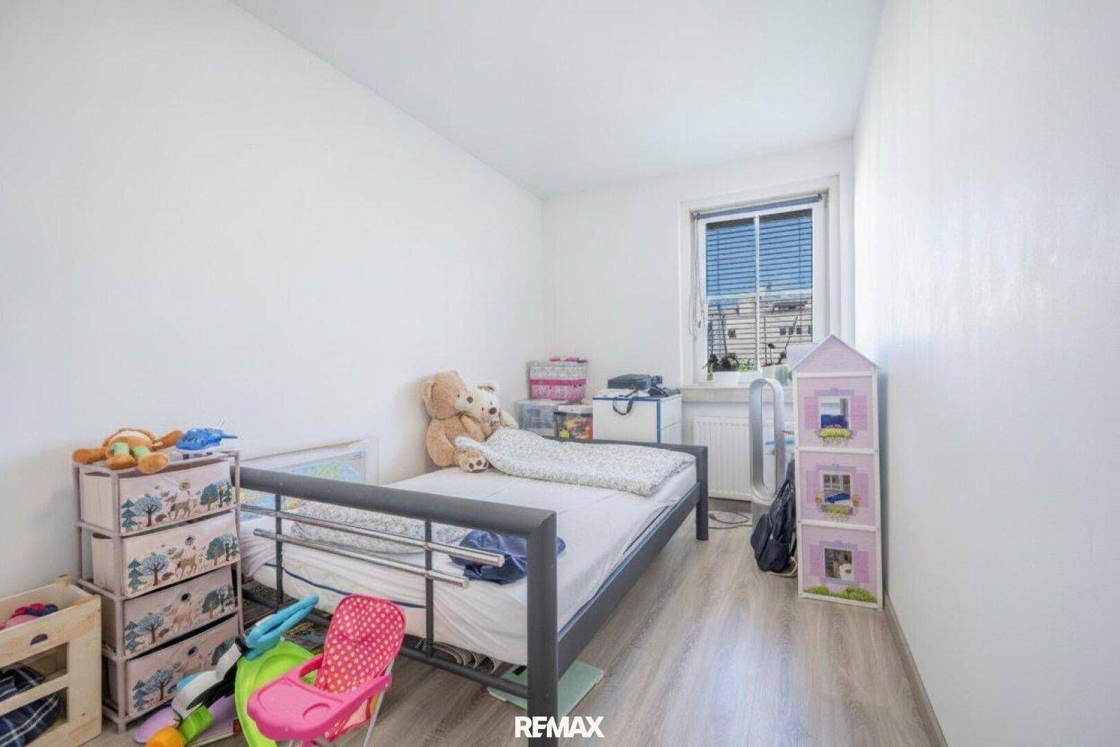 Einfaches Kinderzimmer mit Einzelbett, Spielzeug und einem Fenster mit Jalousien, hell und funktional.