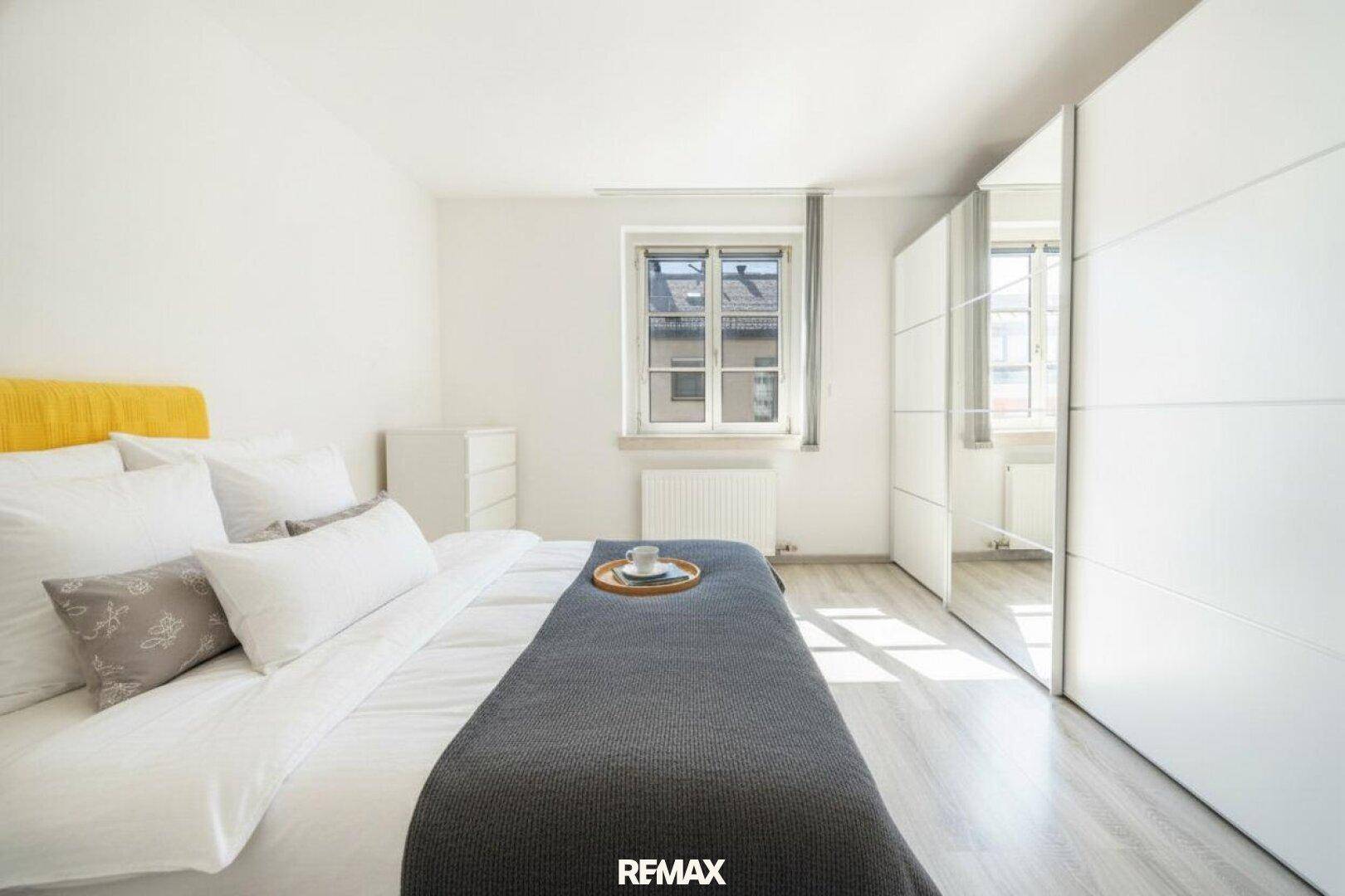 Helles Schlafzimmer mit Doppelbett, weißem Spiegelschrank und Laminatboden, gut beleuchtet durch das Fenster.