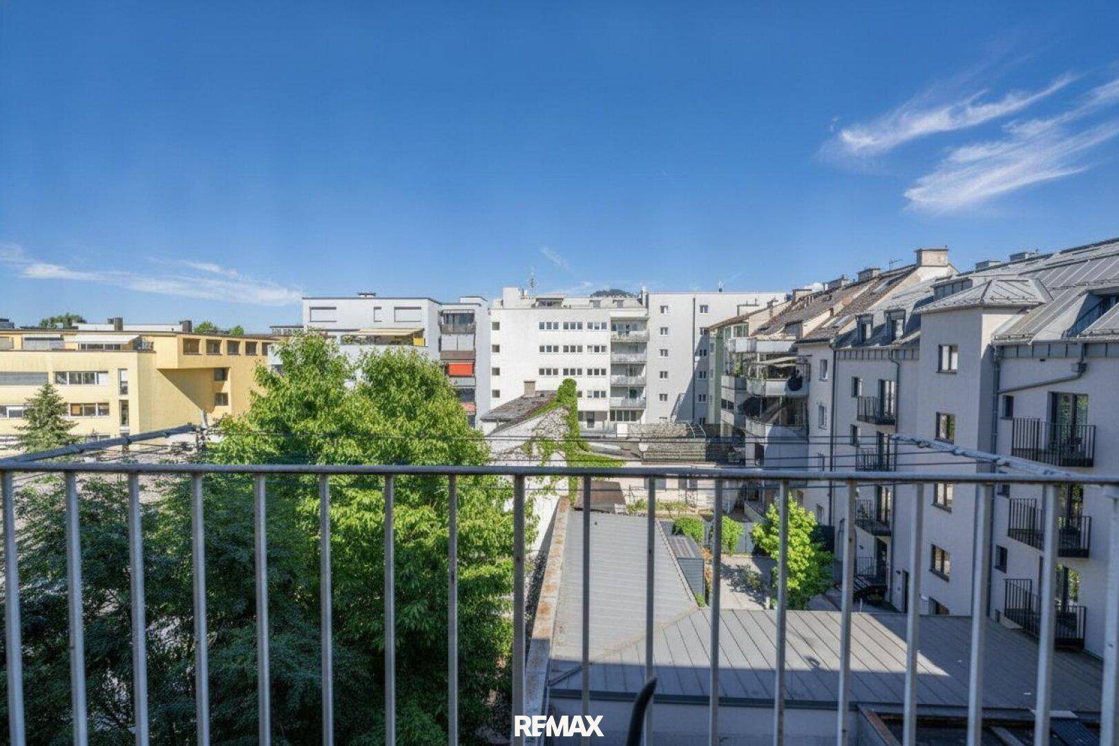 Blick von einem Balkon auf umliegende Gebäude und grüne Bäume unter einem klaren blauen Himmel.