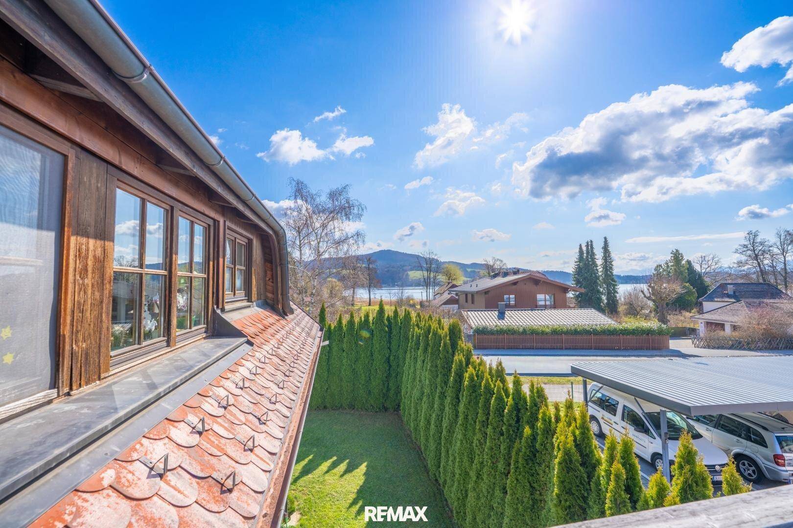 Ausblick auf eine Hecke, einen Carport und den See mit Bergen im Hintergrund an einem sonnigen Tag.