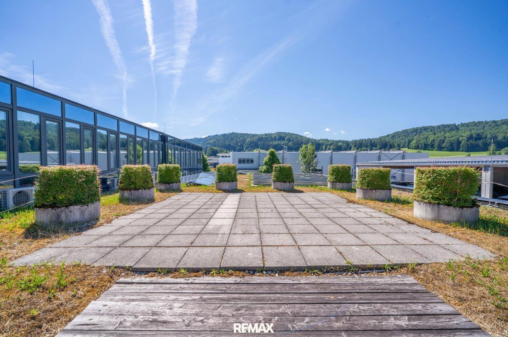 Dachterrasse mit Betonplatten, begrünten Pflanzkübeln und Blick auf die umliegende Landschaft und Gebäude.
