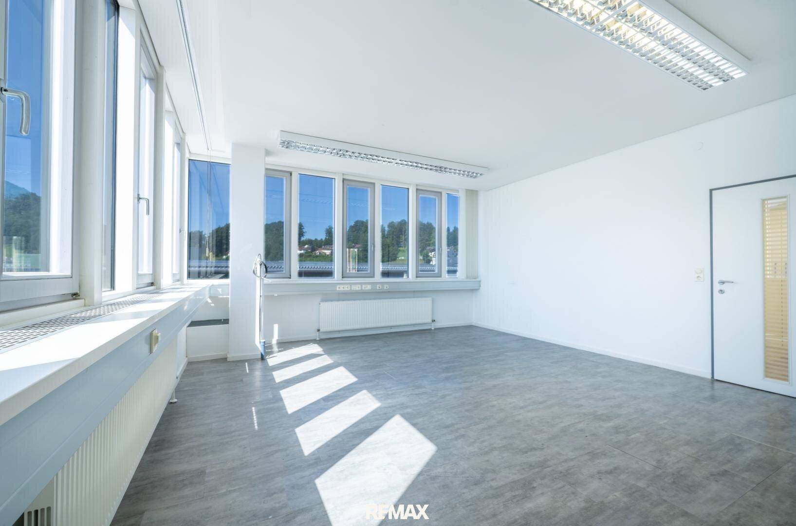 Geräumiger Büroraum mit grauen Fliesen und großen Fenstern, die den Raum mit natürlichem Licht durchfluten.
