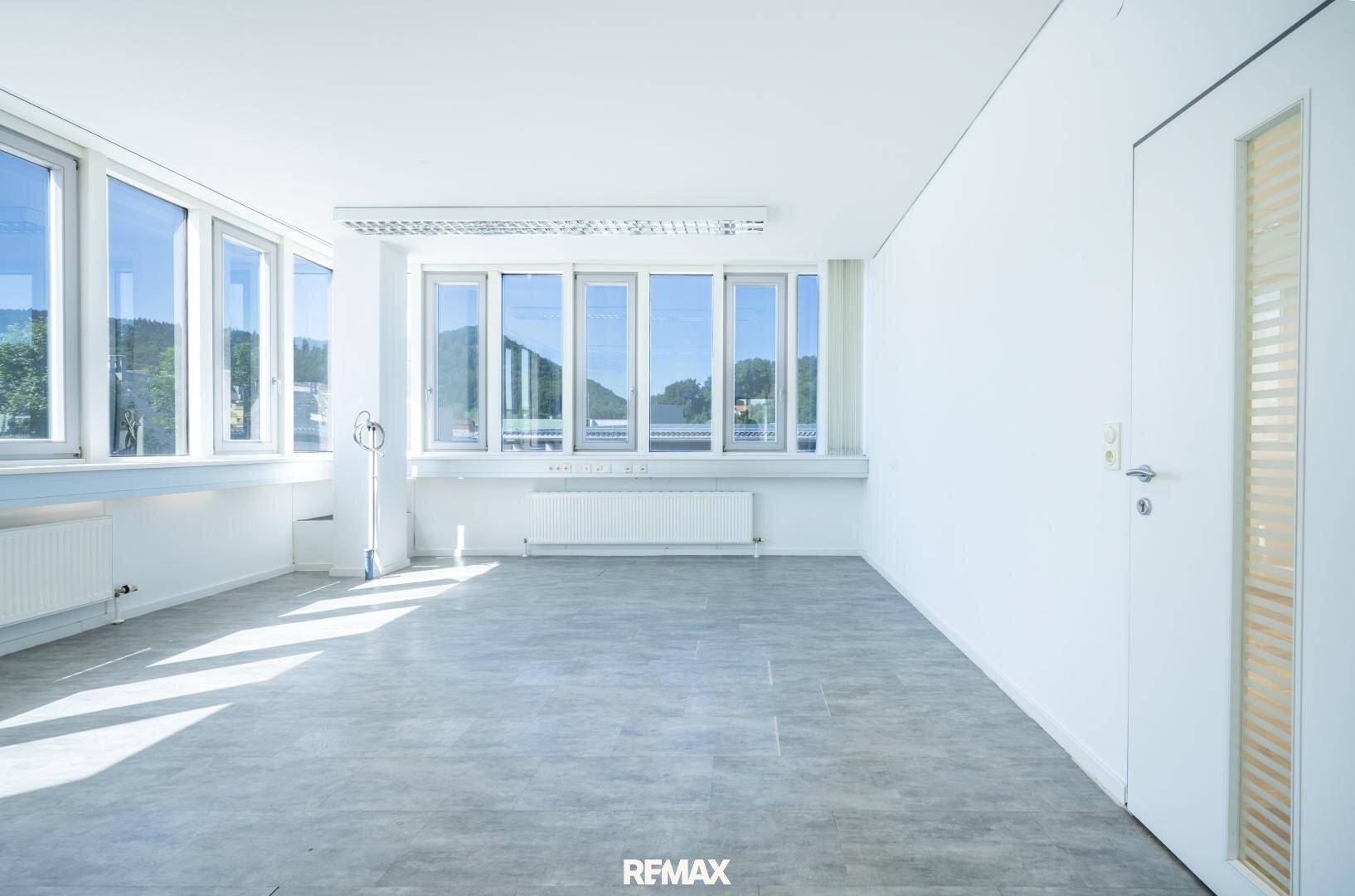Heller, leerer Büroraum mit grauen Fliesen und großen Fenstern, die für eine freundliche Atmosphäre sorgen.