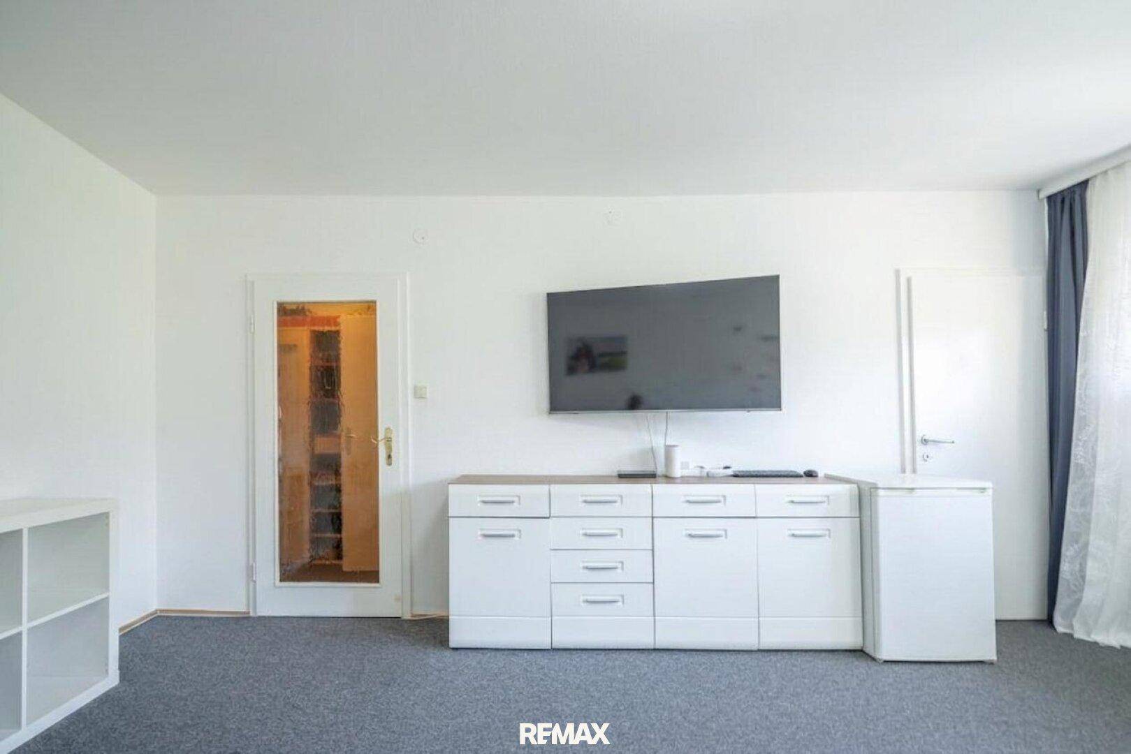 Moderner Wohnbereich mit Wandfernseher, weißem Sideboard und Zugang zu einem Nebenraum.