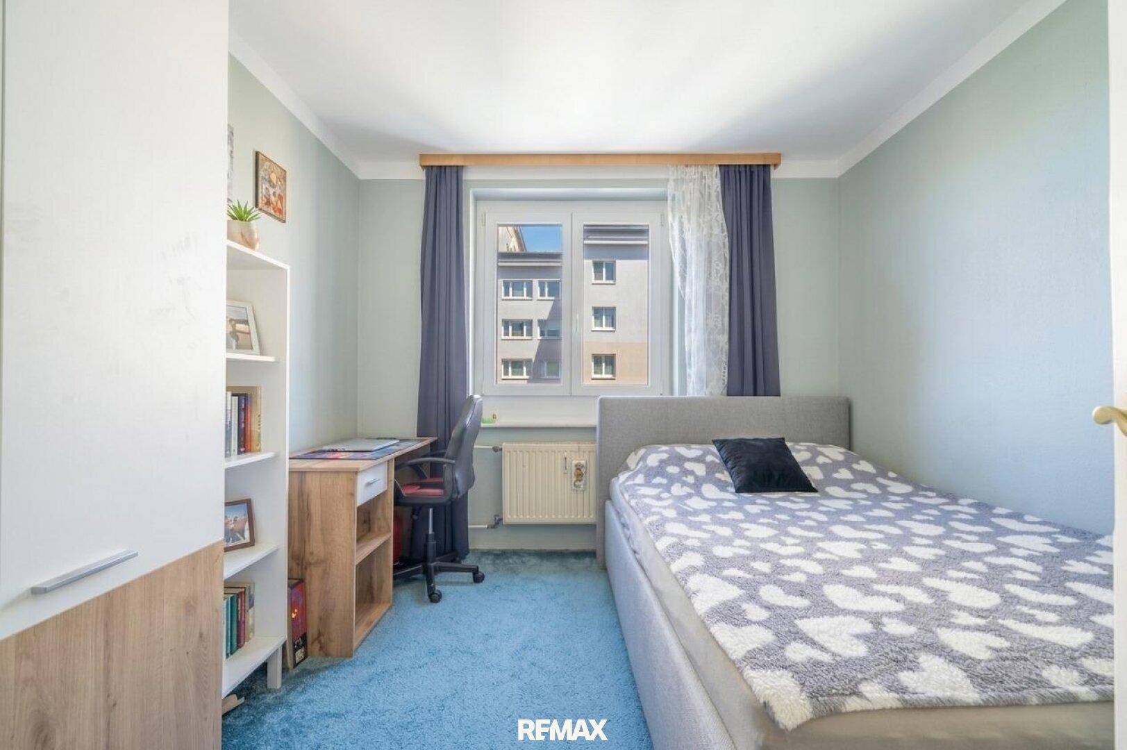 Helles Schlafzimmer mit Einzelbett, Schreibtisch und Blick aus dem Fenster auf die Umgebung.