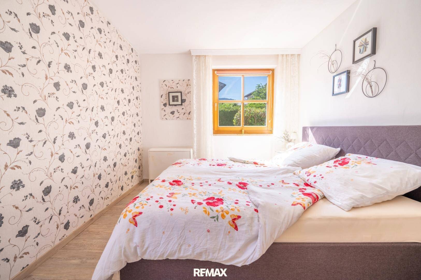 Schlafzimmer mit floraler Tapete, gemütlichem Bett und Fenster mit Blick ins Grüne, sehr hell.