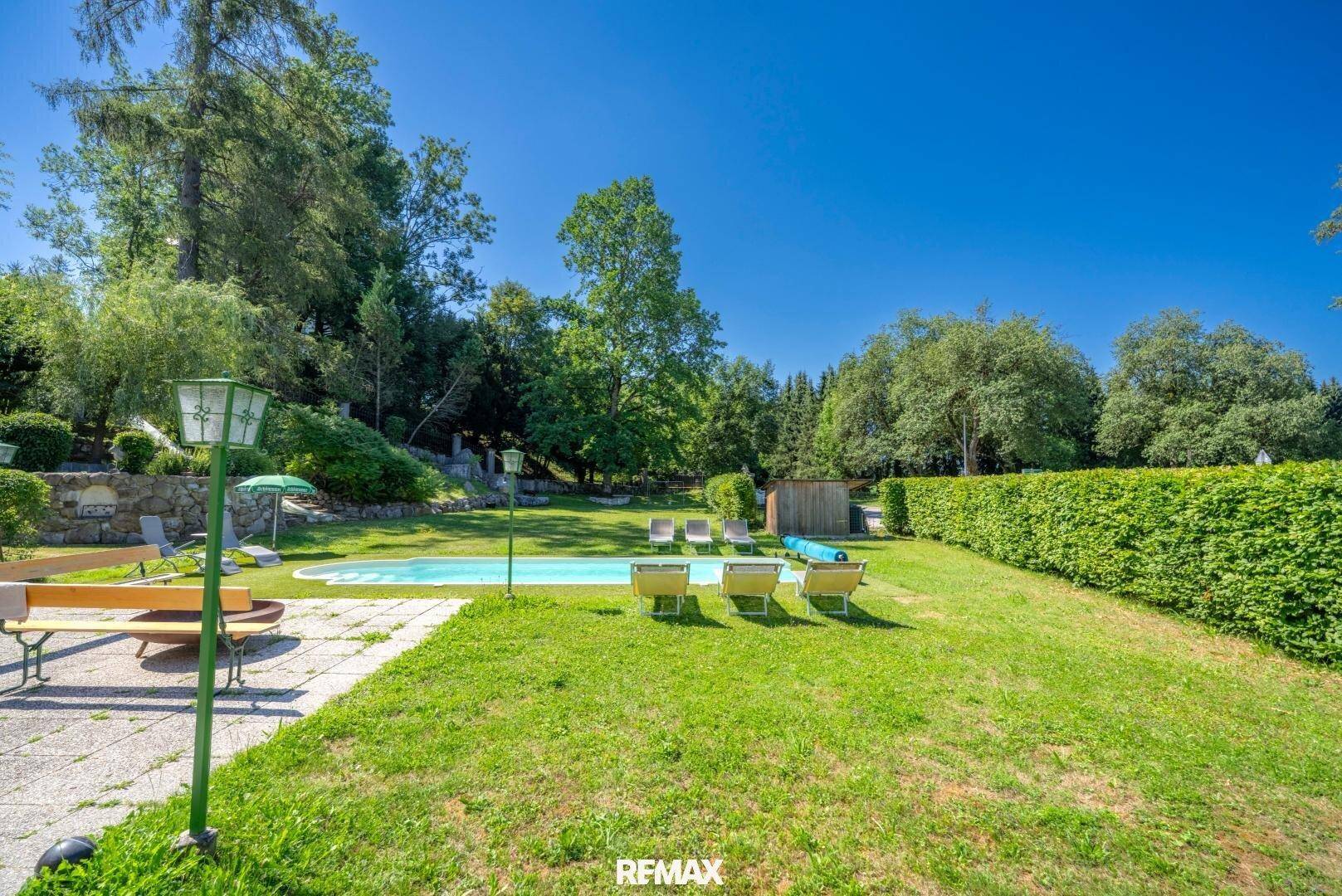 Weitläufiger Garten mit Pool, Liegewiese und hohen Bäumen, umgeben von einer Hecke.