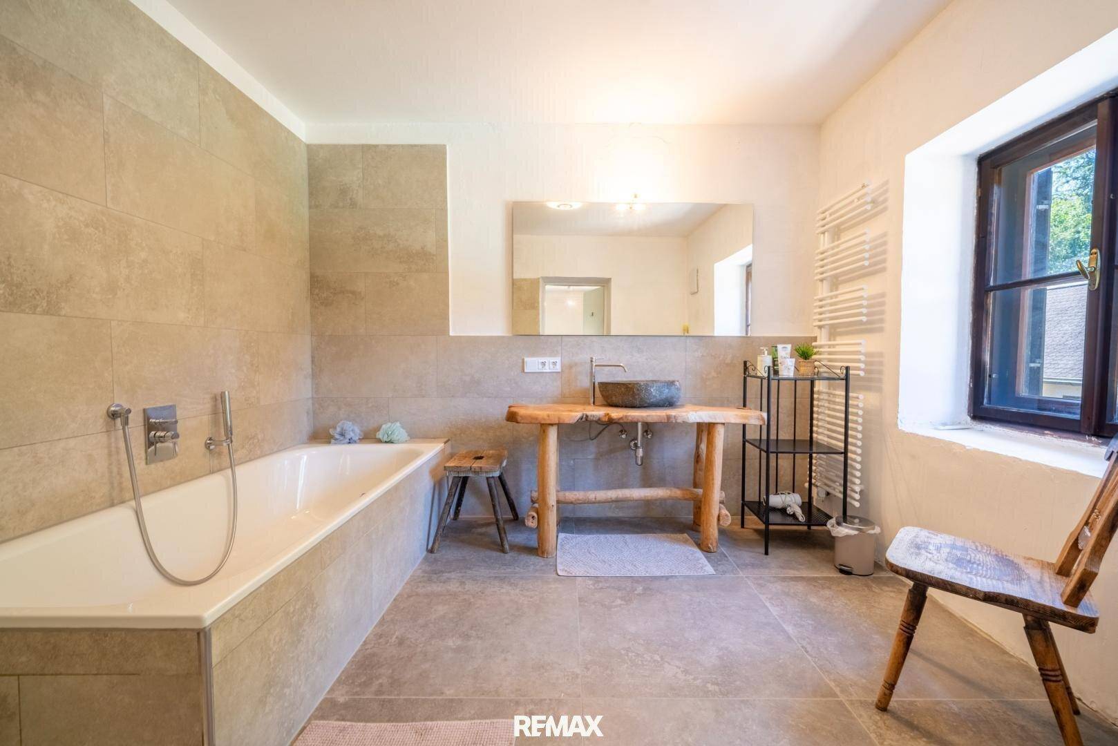 Helles Badezimmer mit Badewanne, rustikalem Waschtisch und Fenster mit Blick ins Grüne.