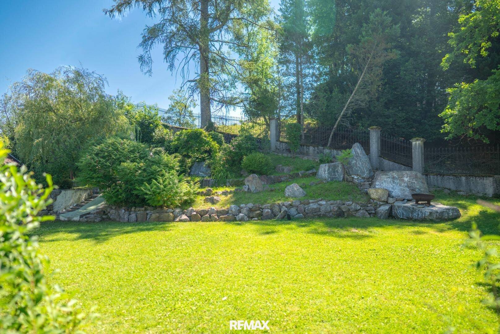 Gepflegter Gartenbereich mit Rasenfläche, Felsen und üppiger Bepflanzung unter blauem Himmel.