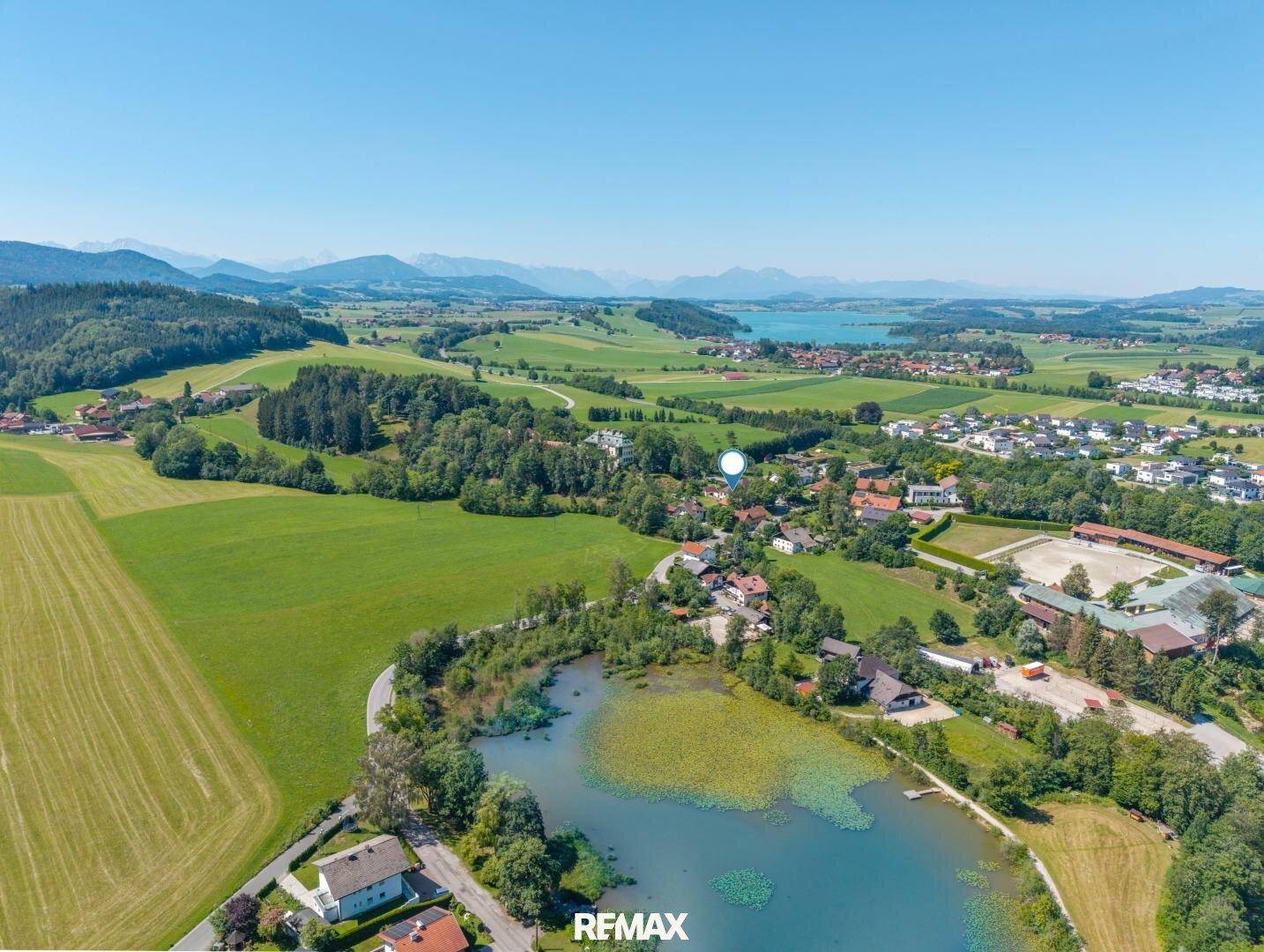 Weitläufige Luftaufnahme einer malerischen Landschaft mit See, grünen Feldern und verstreuten Häusern unter klarem Himmel.