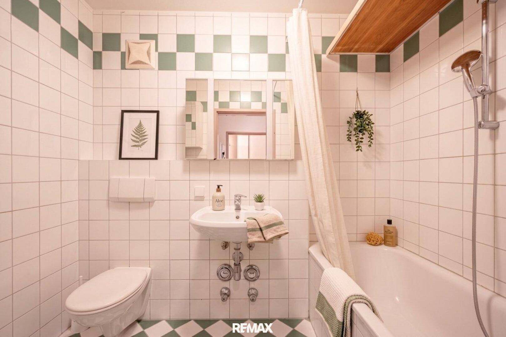 Badezimmer mit weiß-grünen Fliesen, Badewanne mit Duschvorhang und Spiegelschrank über dem Waschbecken.