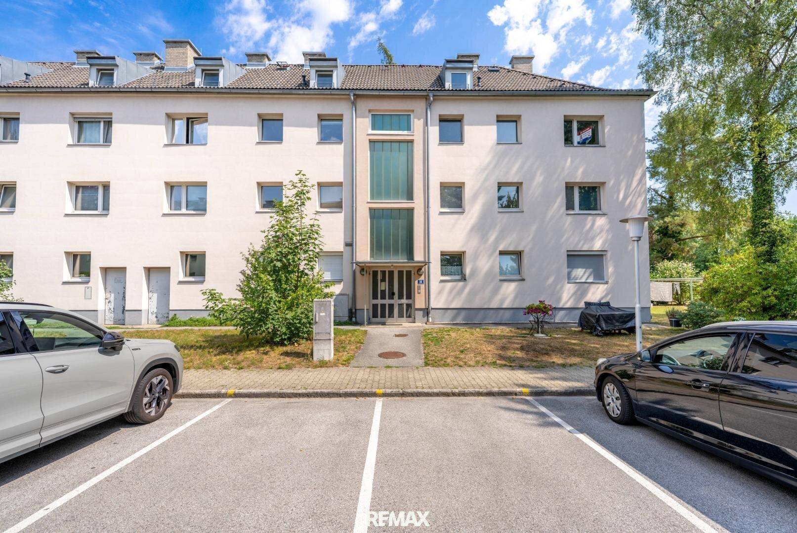 Mehrfamilienhaus mit heller Fassade, vielen Fenstern und Parkplätzen im Vordergrund.