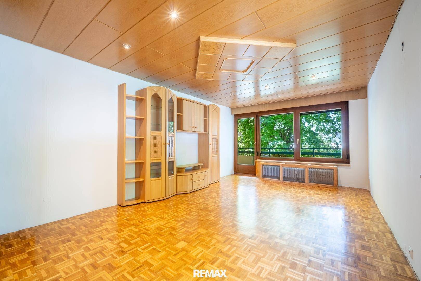 Geräumiges Wohnzimmer mit Holzdecke, Parkettboden und großem Fenster mit Blick ins Grüne.