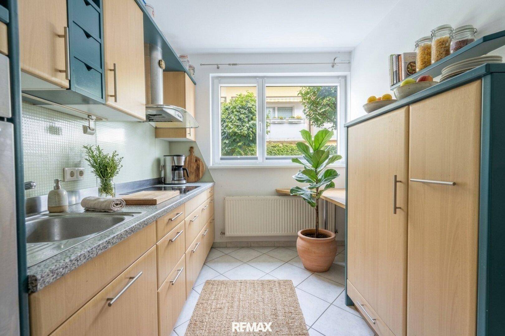 Funktionale Küche mit hellen Holzschränken, Fliesenboden und Fenster mit Blick nach draußen.