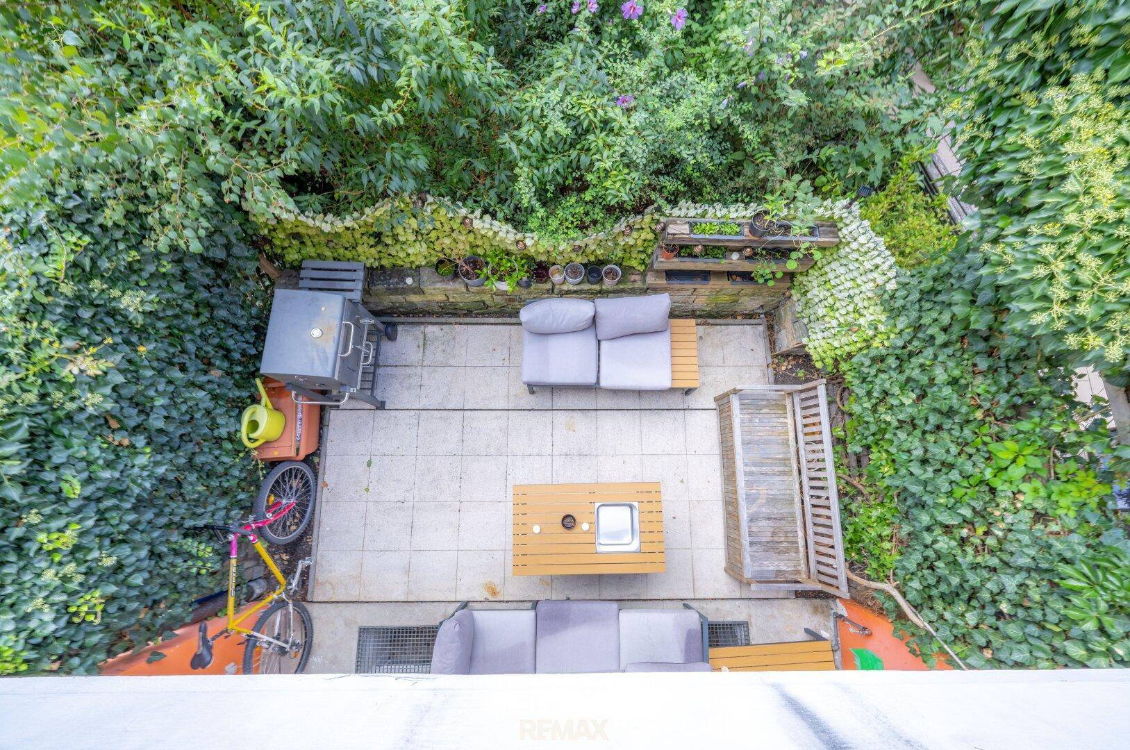 Vogelperspektive auf die begrünte Terrasse mit Sitzgelegenheiten und Grill, umgeben von Pflanzen.