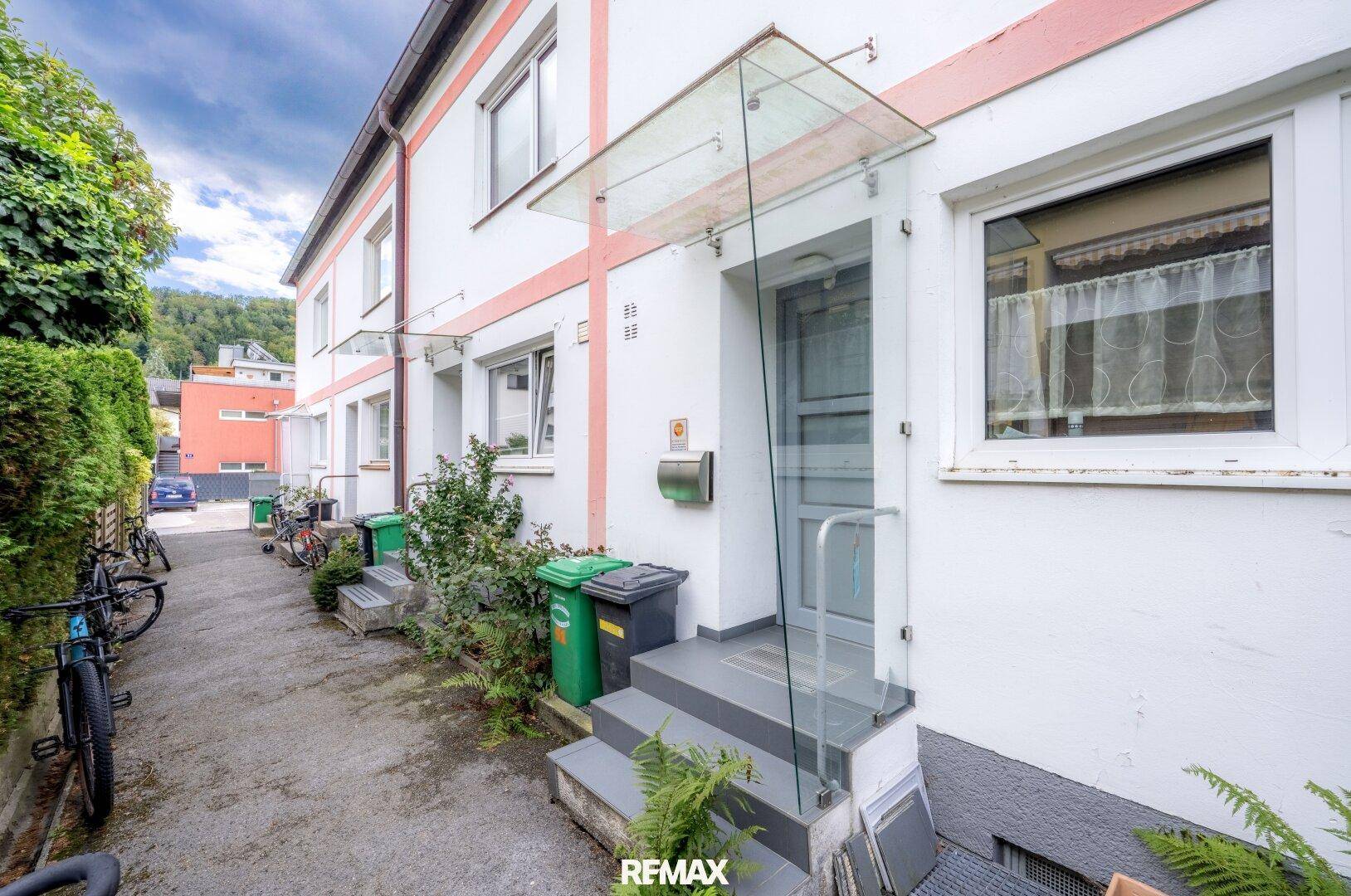 Eingangsbereich eines Reihenhauses mit Vordach und Zugang zur Straße, umgeben von Grün.