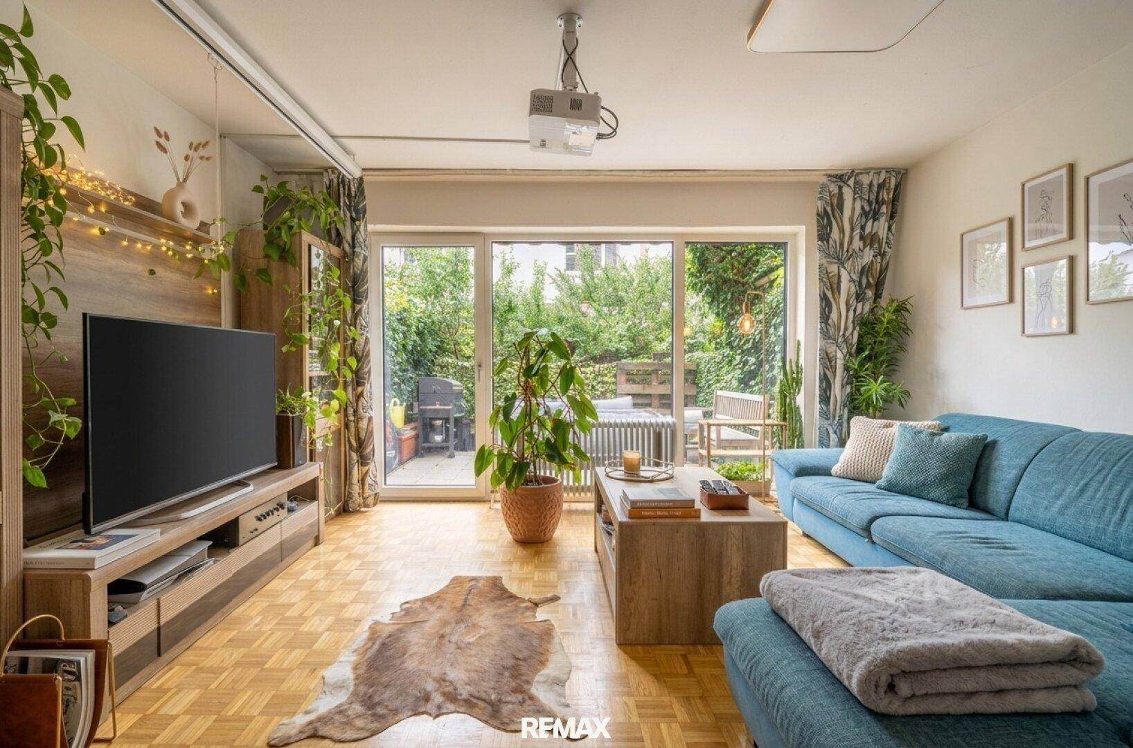 Geräumiges Wohnzimmer mit Holzboden, großem Fernseher und Zugang zur grünen Außenterrasse.