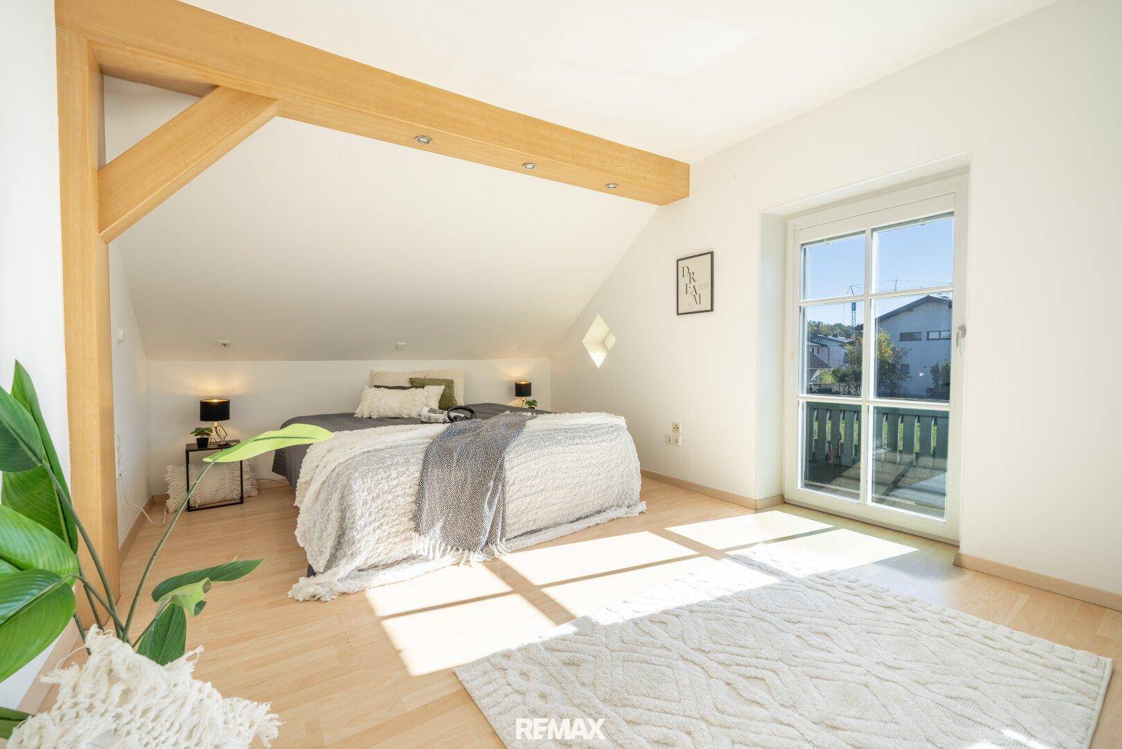 Schlafzimmer im Dachgeschoss mit Holzbalken, schräger Decke und Zugang zu einem französischen Balkon.