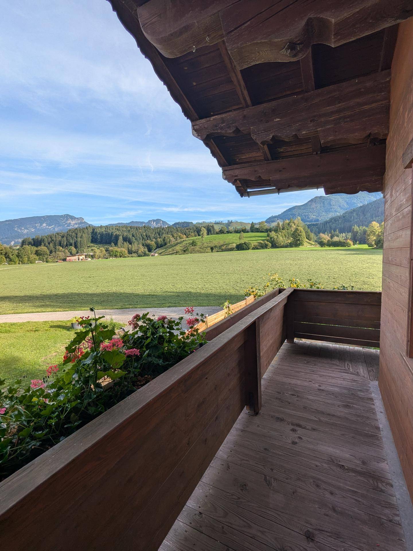 Weiter Blick vom Holzbalkon über weite grüne Felder und die beeindruckende Bergkulisse.