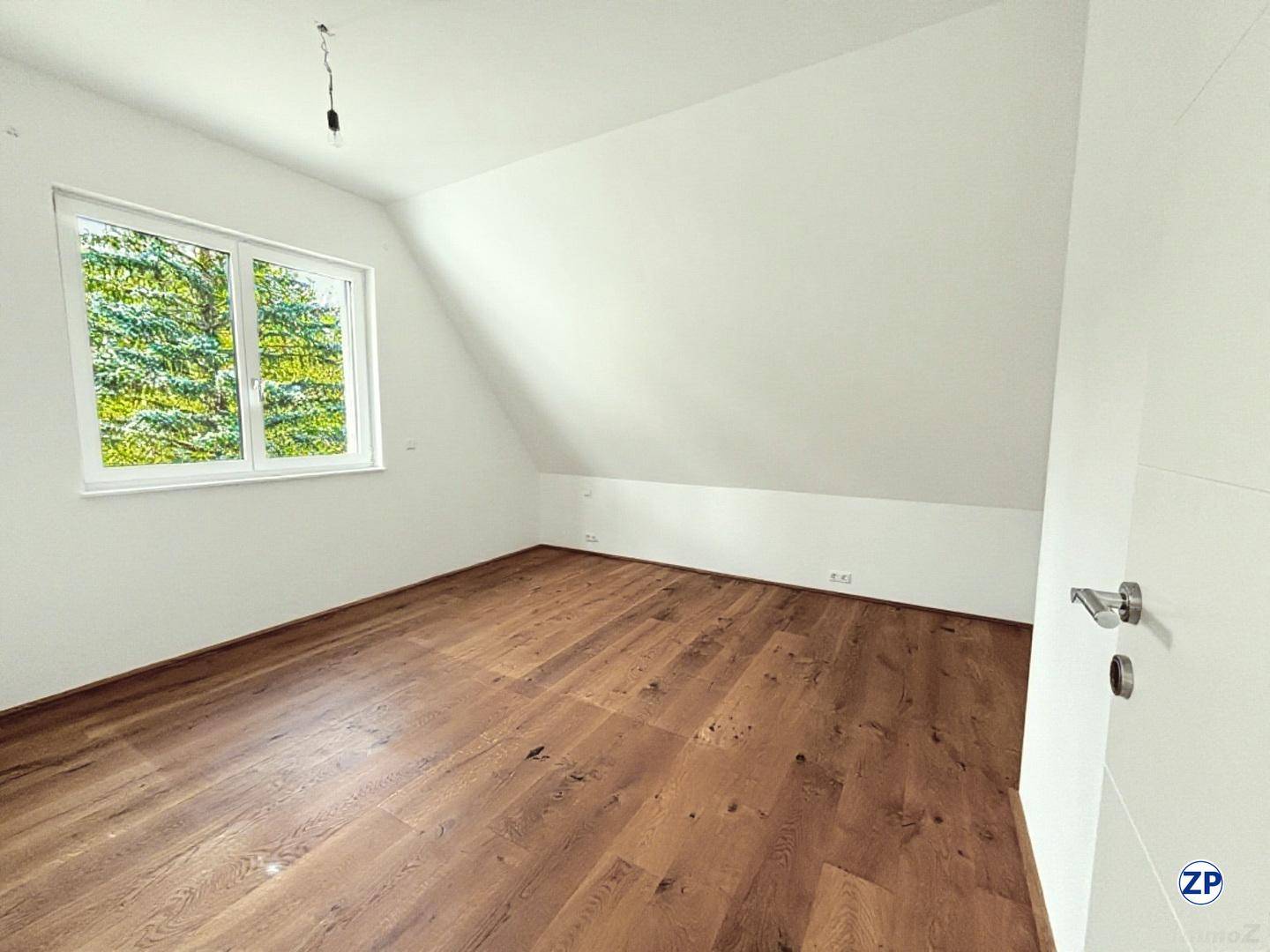 Helles Schlafzimmer im Dachgeschoss mit Holzboden und einem Fenster mit Blick ins Grüne.