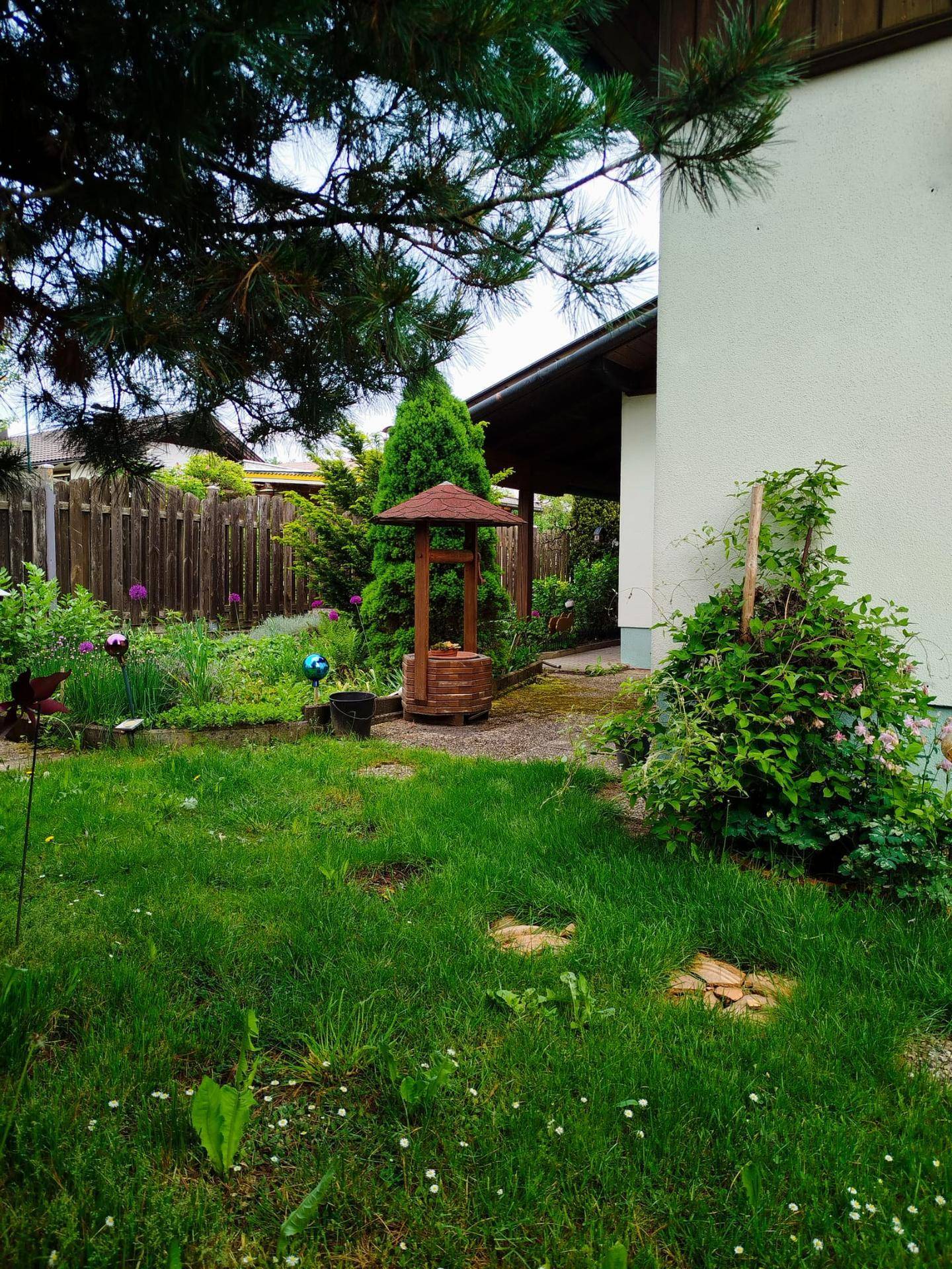 Gepflegte Rasenfläche im Garten mit einem dekorativen Holzbrunnen und umgebenden Pflanzen unter Bäumen.