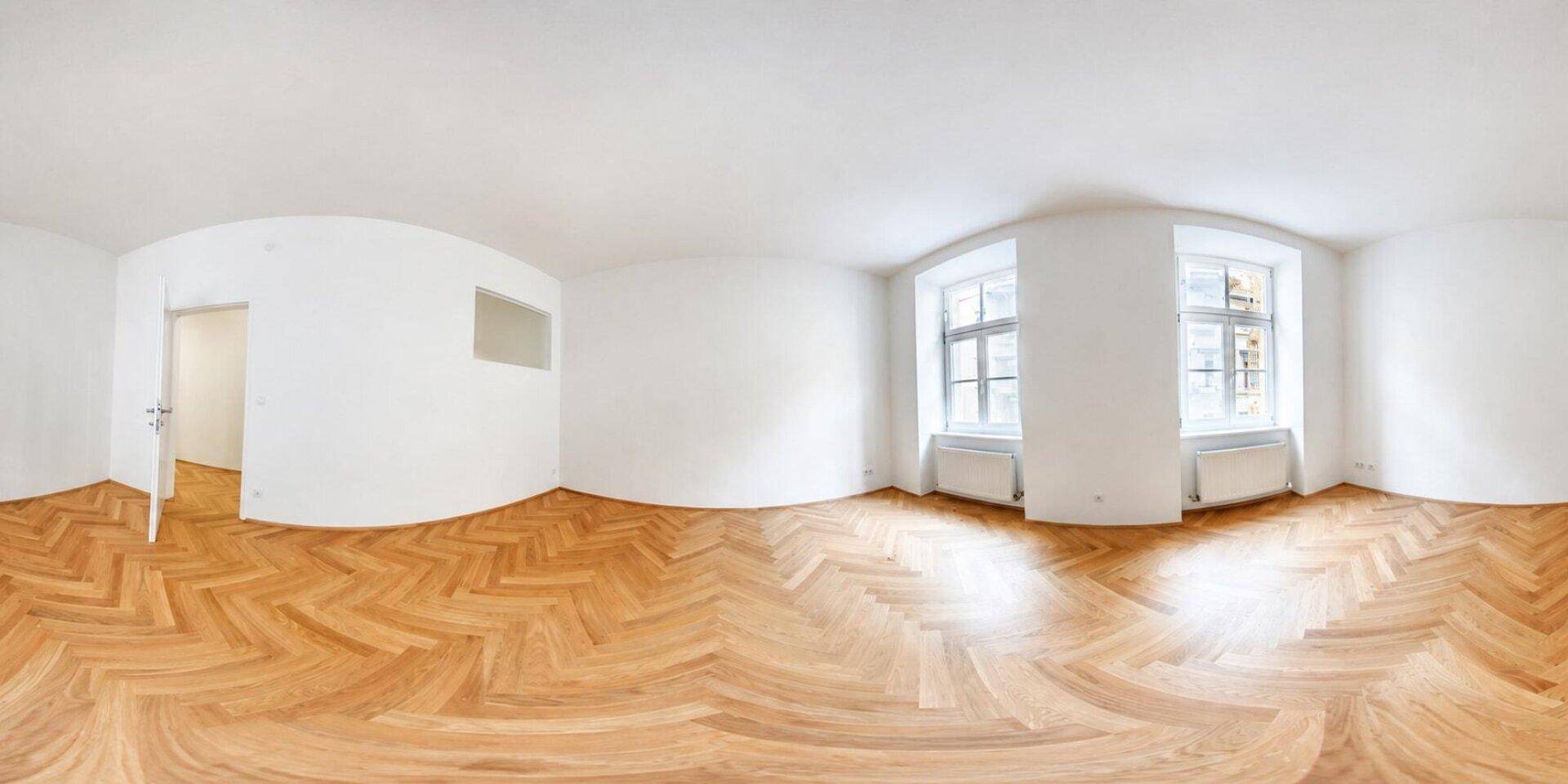 Heller, leerer Raum mit Fischgrätparkett und zwei Fenstern, die viel Tageslicht hereinlassen.