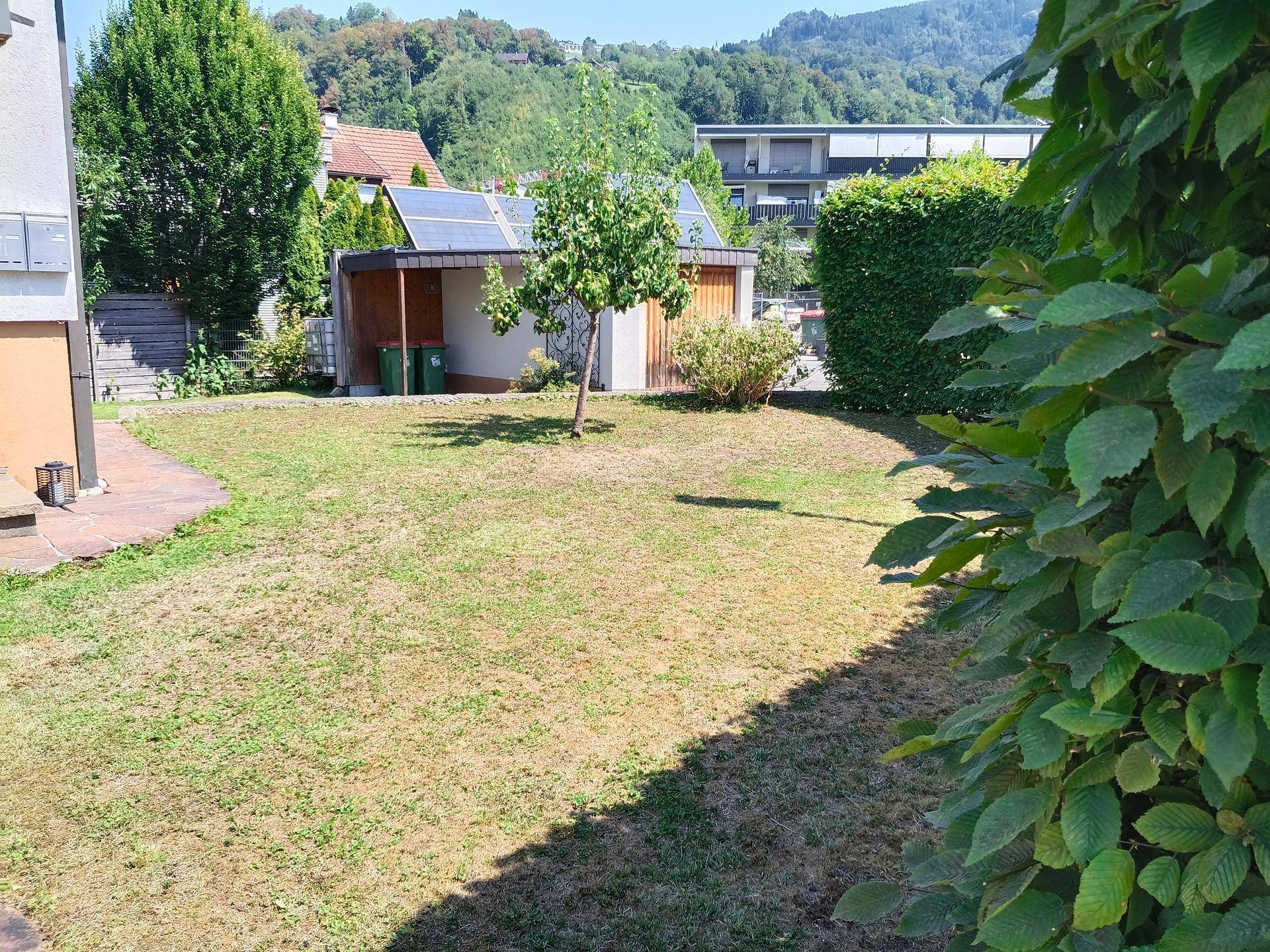 Gepflegter Garten mit Rasenfläche, Bäumen und Hecke, im Hintergrund sind grüne Hügel sichtbar.
