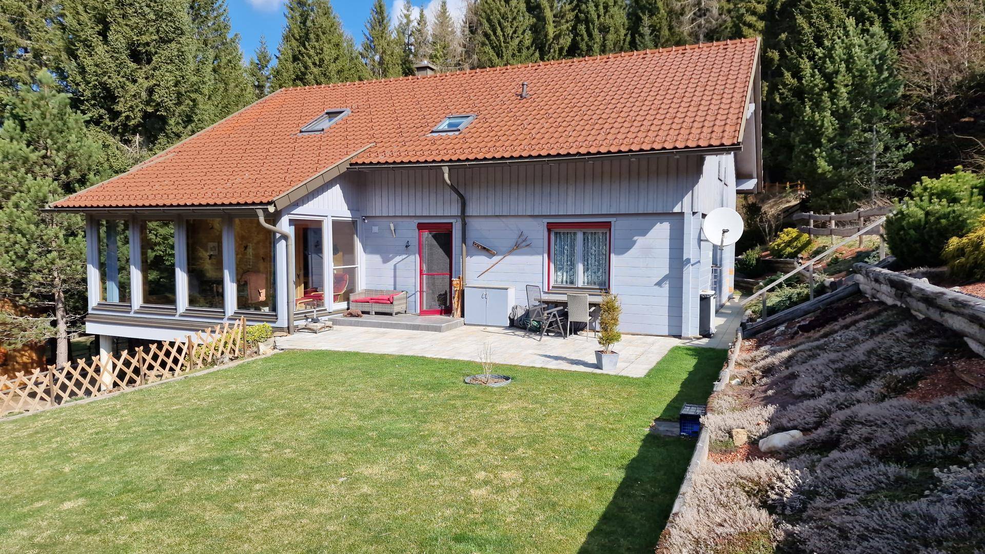 Rückansicht des Hauses mit gepflegtem Rasen, Terrasse und Dachfenstern, umgeben von Natur.