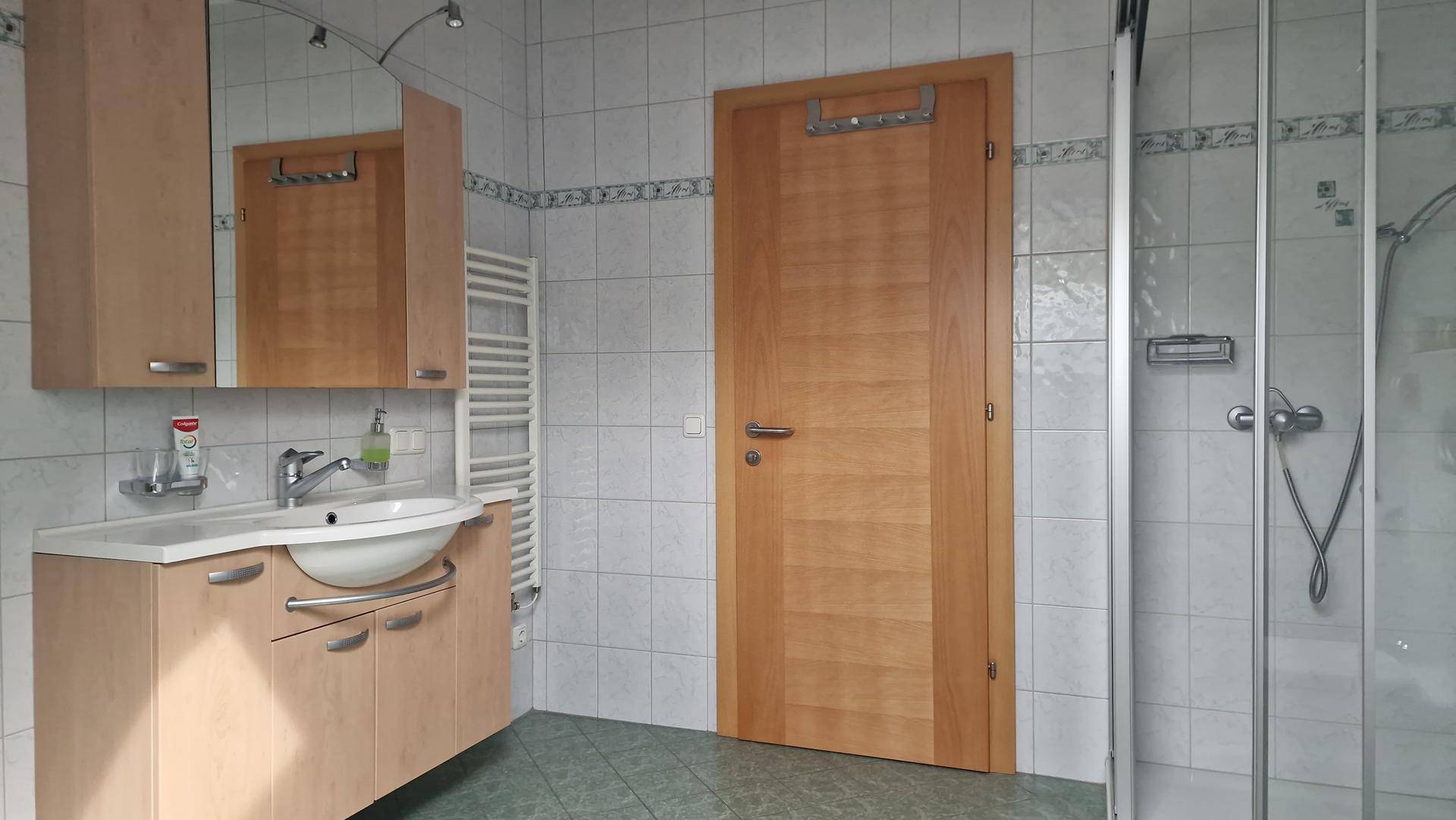 Badezimmer mit Duschkabine, hellem Holzwaschtisch und weiß-grauen Fliesen an den Wänden.