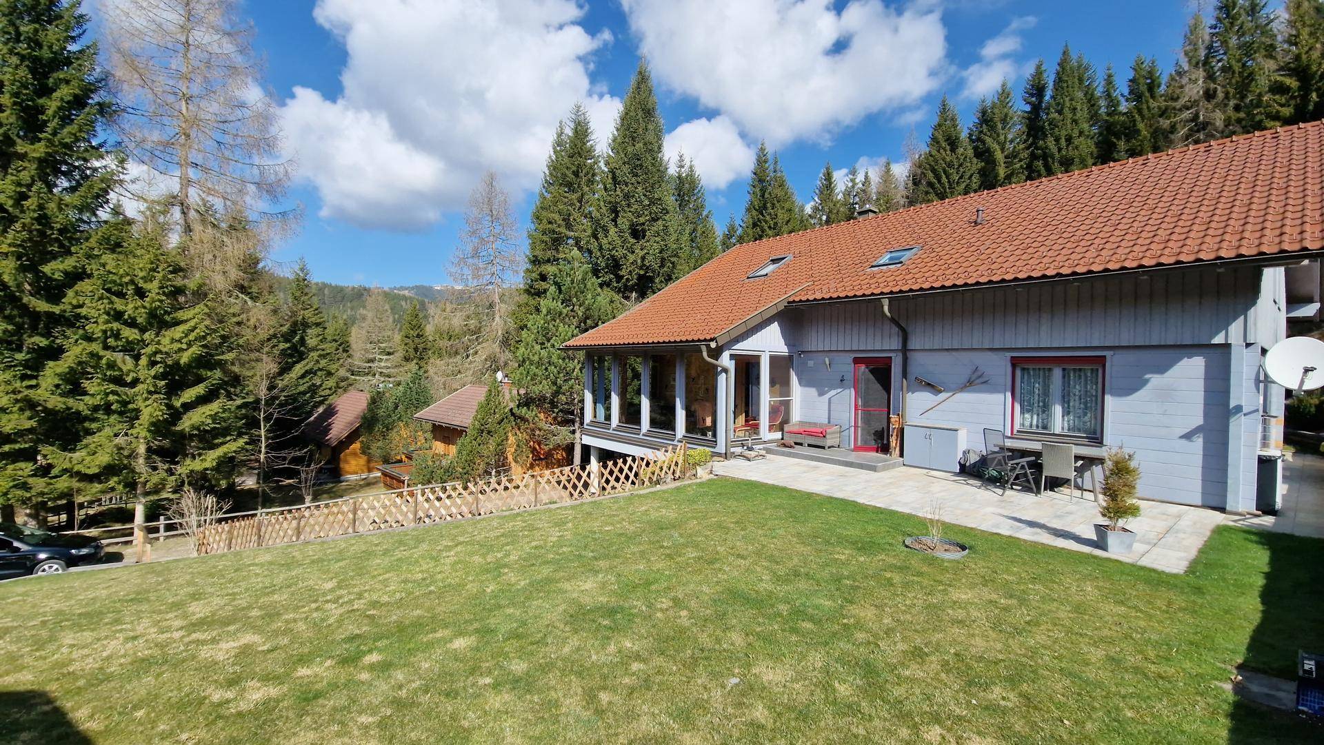 Hinterer Gartenbereich mit großer Rasenfläche und gepflasterter Terrasse, umgeben von hohen Bäumen.