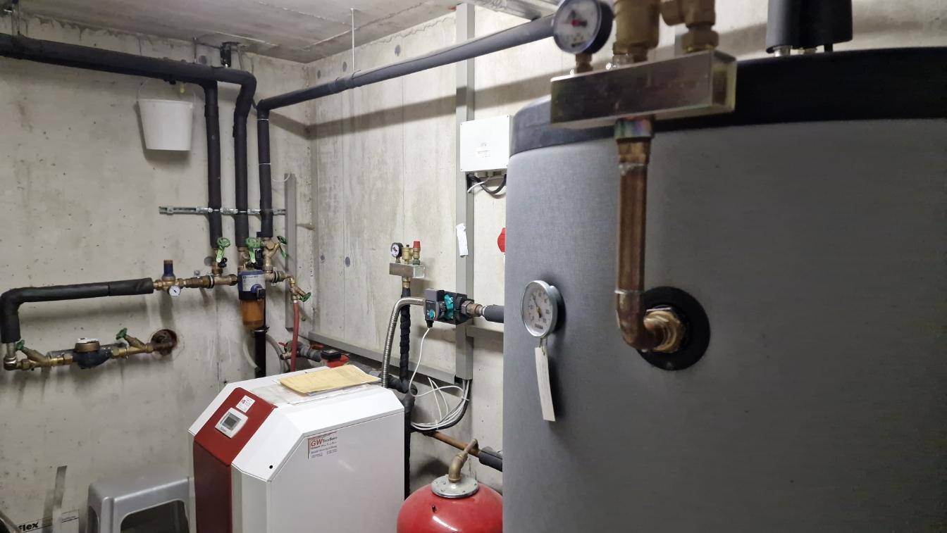 Heizungsraum mit Warmwasserspeicher und komplexer Rohrinstallation an der Wand.