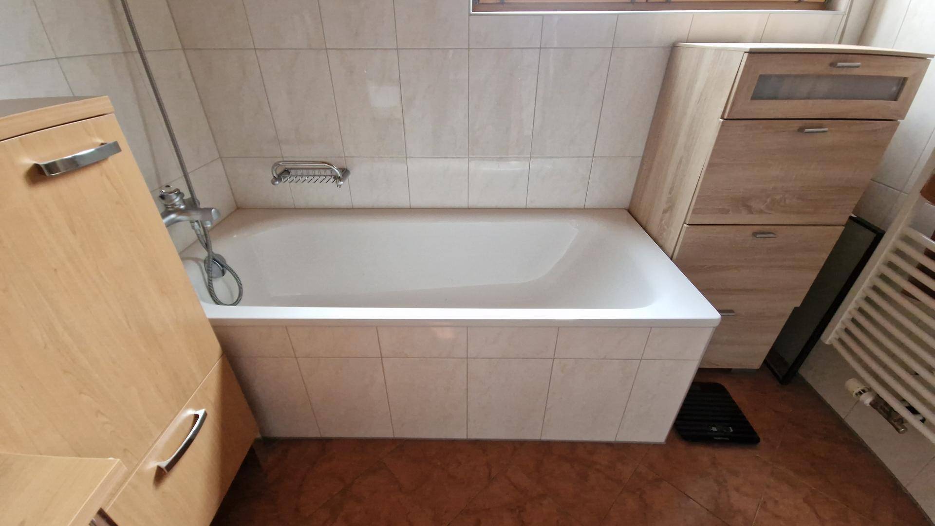 Badezimmer mit Badewanne, hellen Fliesen und Holzschränken für Stauraum.