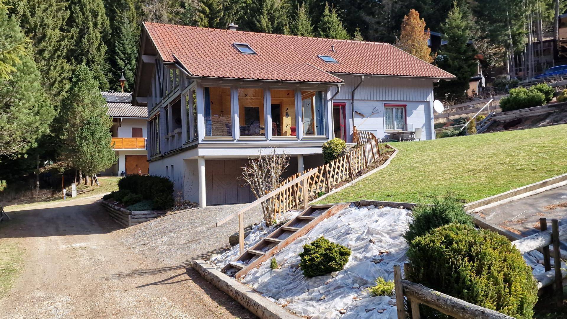 Haus mit rotem Ziegeldach und Hanglage, umgeben von grüner Landschaft und Bäumen.