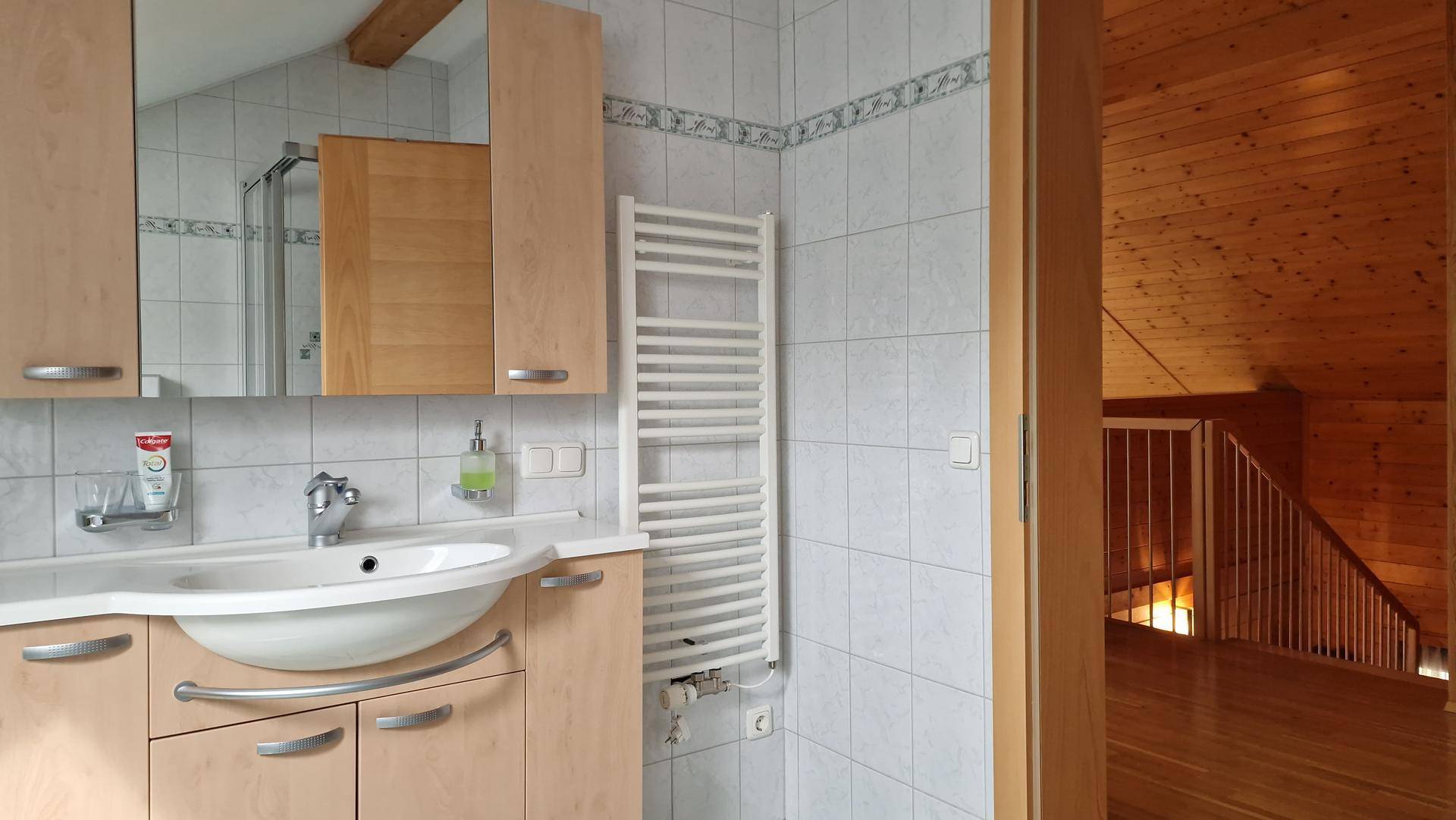 Badezimmer mit hellem Holzwaschtisch, Spiegel und Handtuchheizkörper, angrenzend an Flur.