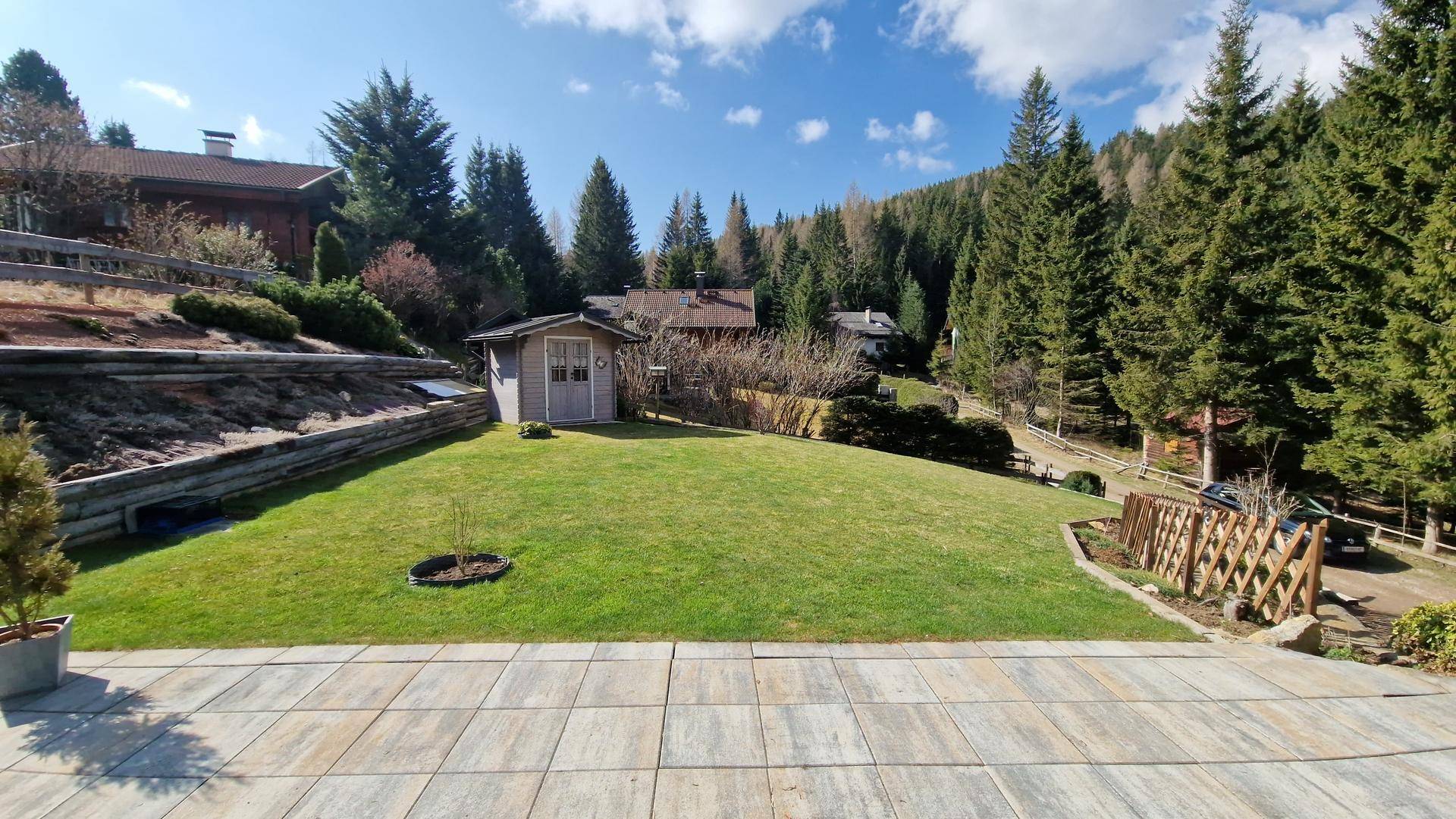 Sonniger Gartenbereich mit gepflasterter Terrasse, Rasenfläche und einem kleinen Gartenhaus vor Waldkulisse.