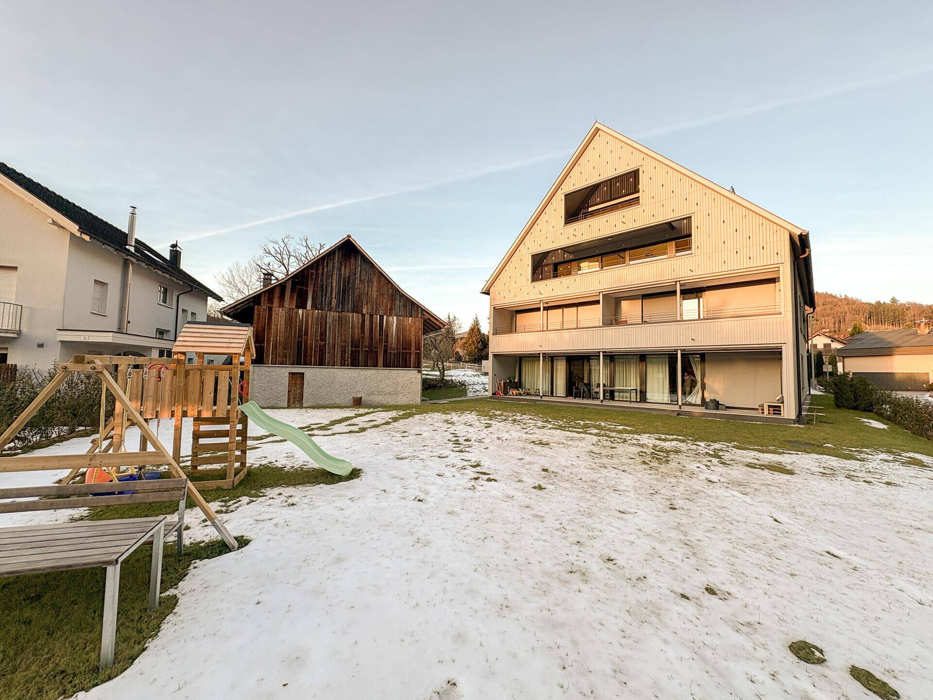 Hinterhof mit Schnee, modernem Gebäude und einem Kinderspielplatz in ländlicher Umgebung.