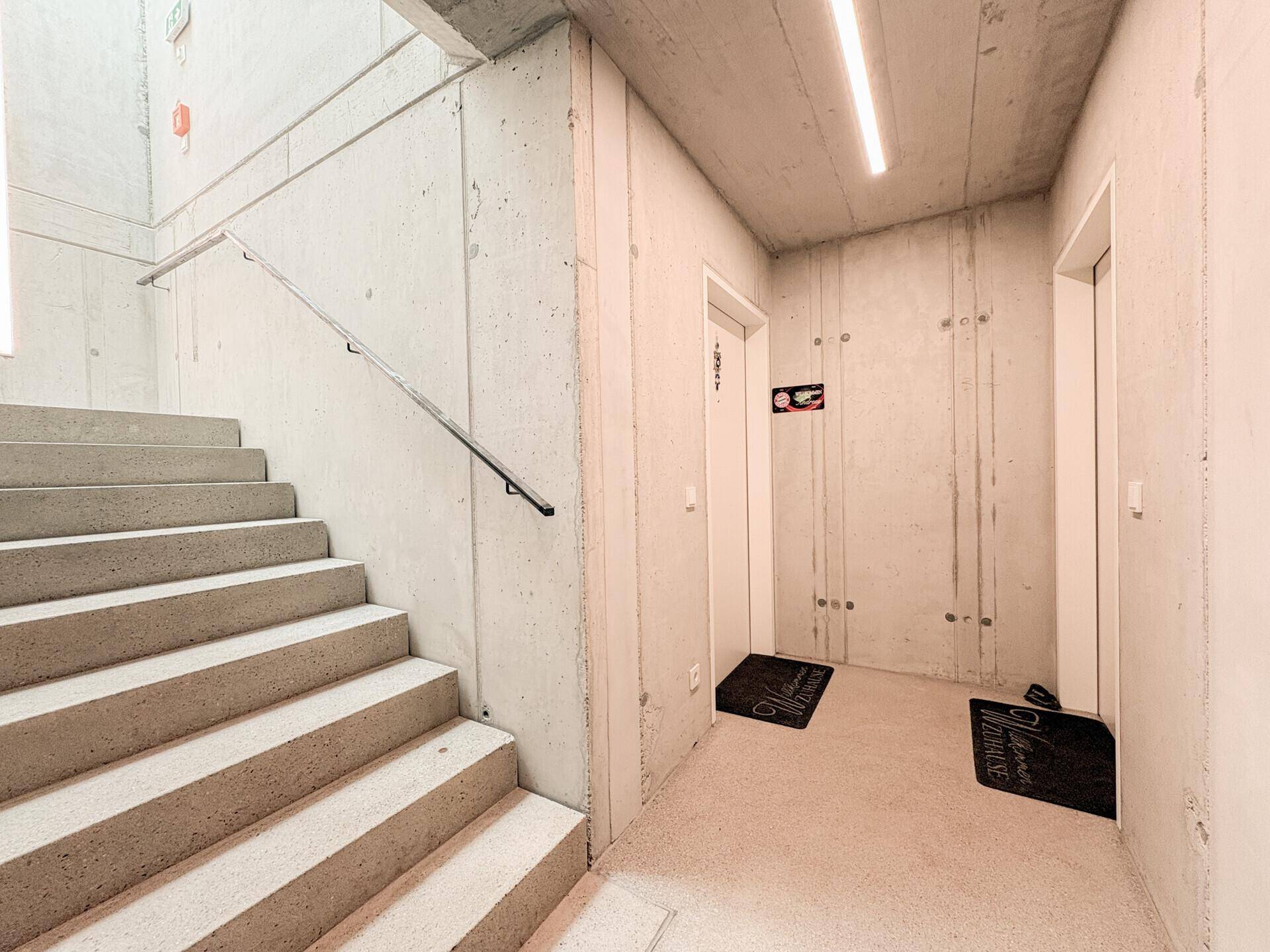 Heller Flur mit Betonwänden, Treppenaufgang und Zugang zu den Wohneinheiten.